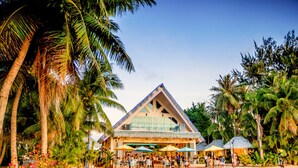 A beachside building with a triangular roof, surrounded by palm trees and a sandy beach. - Surfrider Resort Hotel (Saipan)