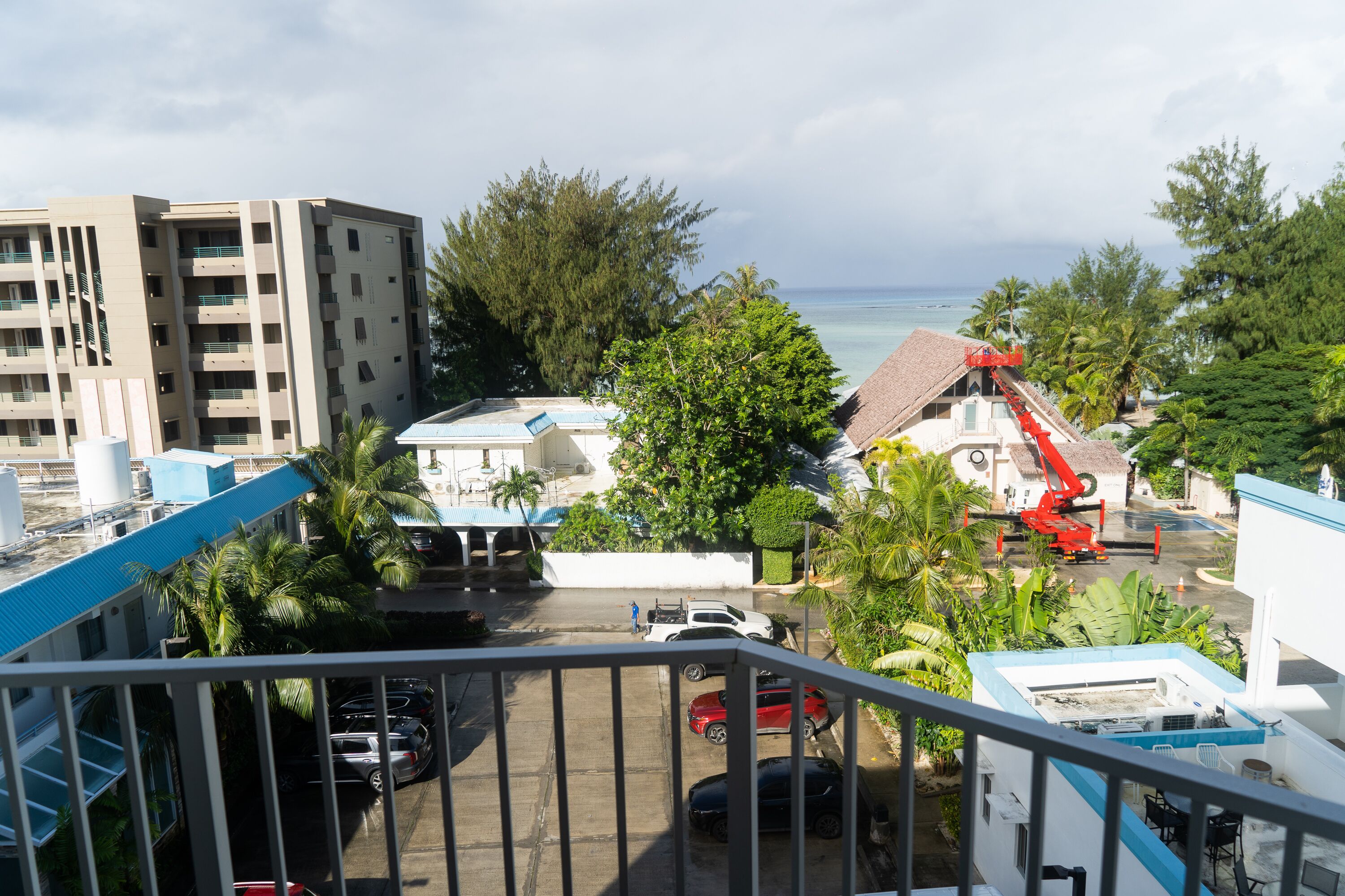 A view from a balcony overlooking a parking area with several cars, a red crane, and a building with a red roof.