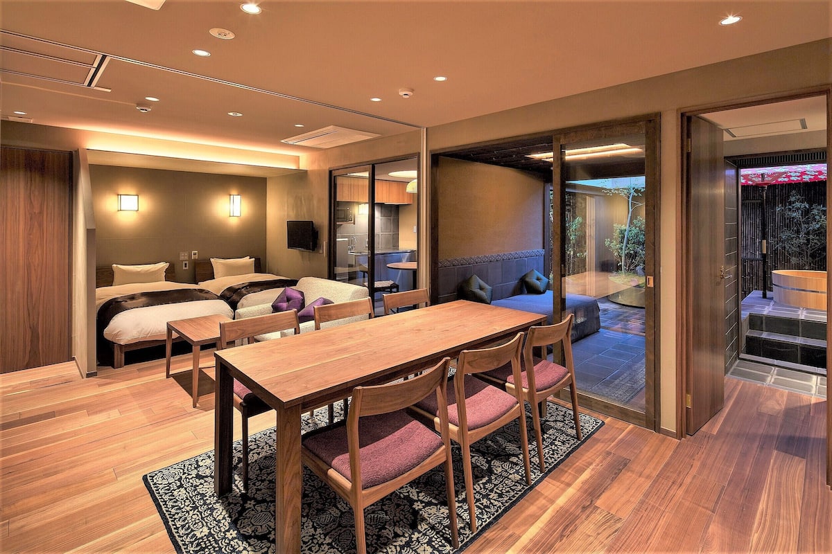 A modern hotel room with a wooden dining table, cushioned chairs, a sofa, and a large window.