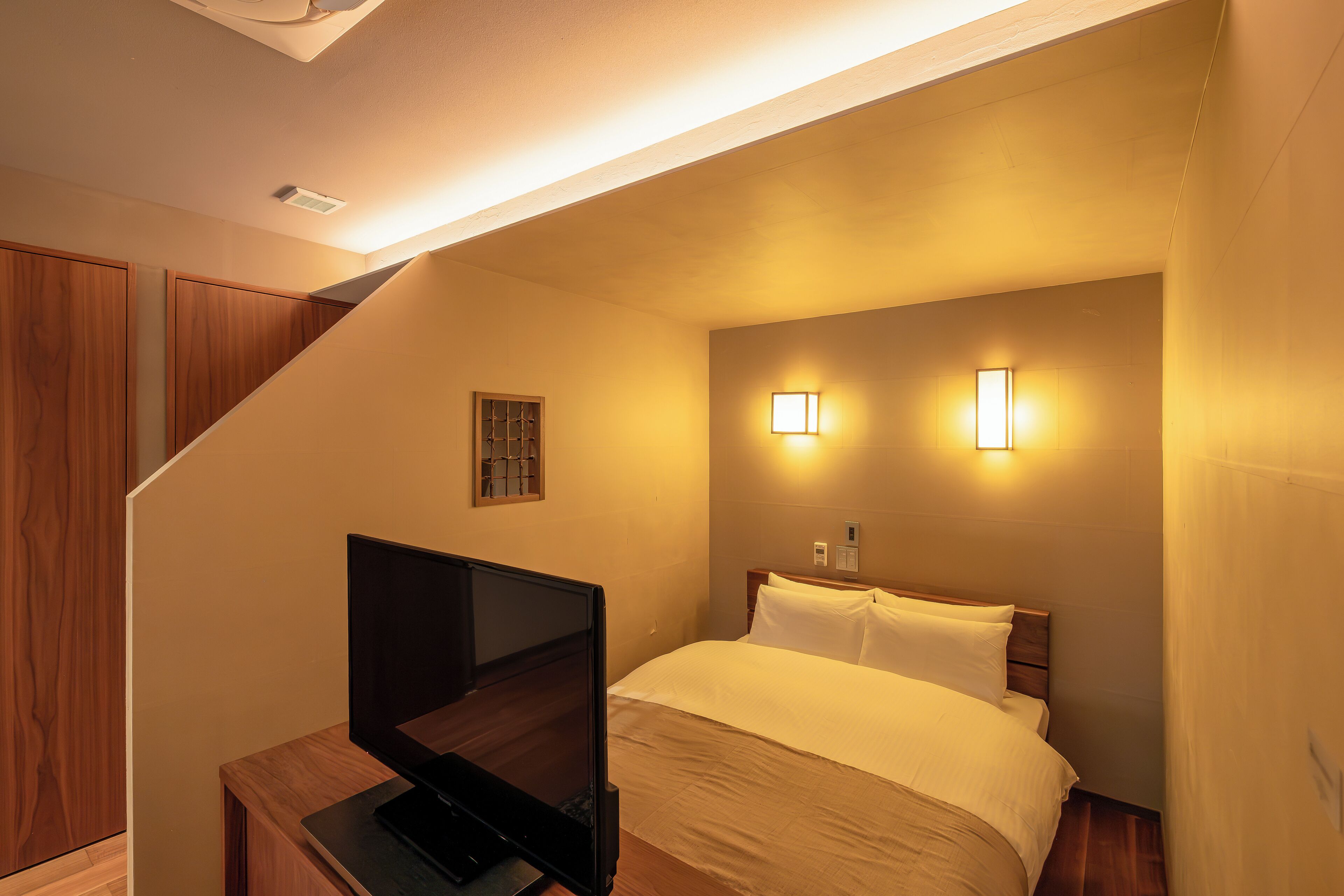 Une chambre d’hôtel avec un lit, un téléviseur posé sur un meuble en bois et deux lampes fixées au mur.