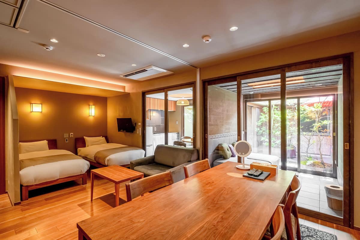 A hotel room with a wooden dining table, two beds, a sofa, and a sliding glass door leading to a balcony with plants.