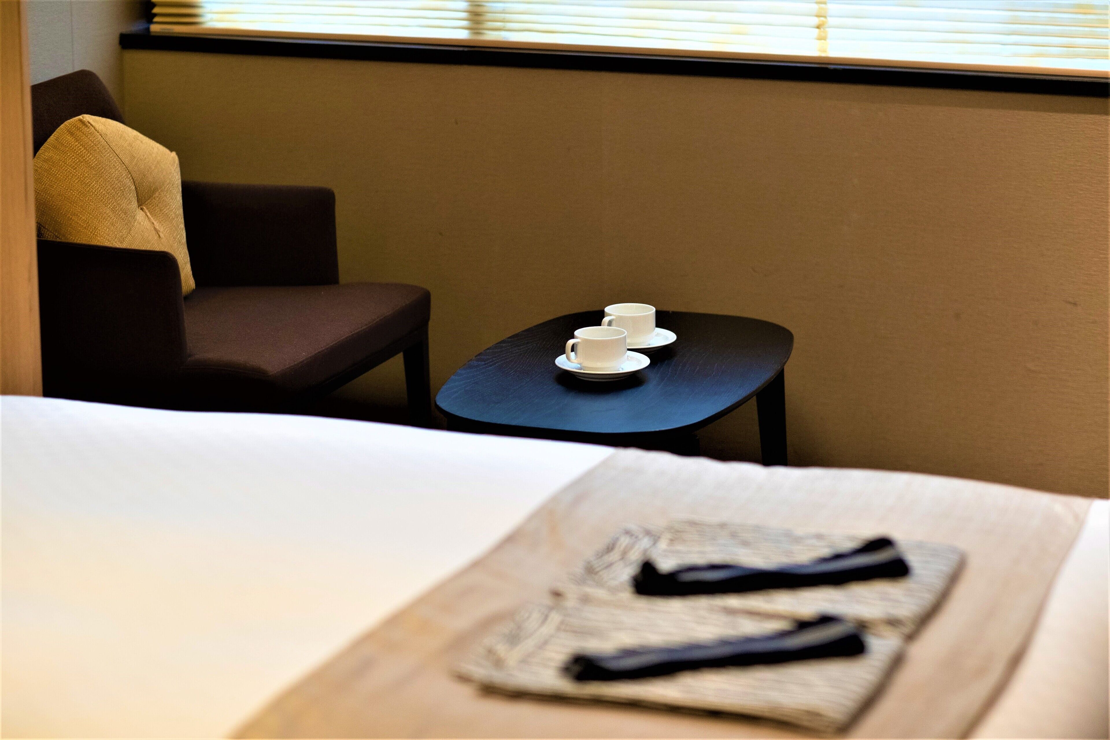 A hotel room with a bed, a small table with cups, a chair, and a window with blinds.