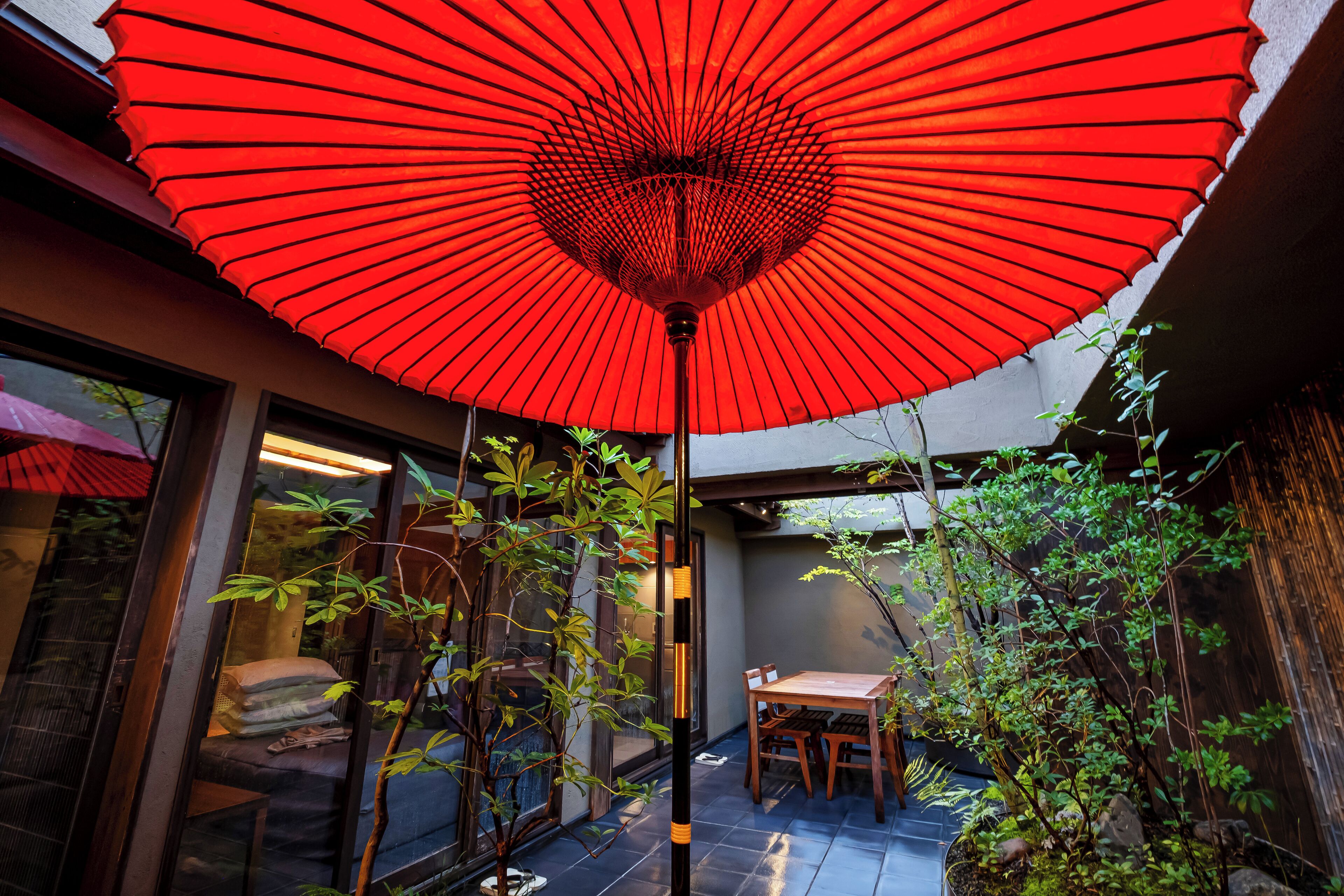 A red umbrella in a courtyard with a table and chairs, surrounded by plants and a wooden fence.