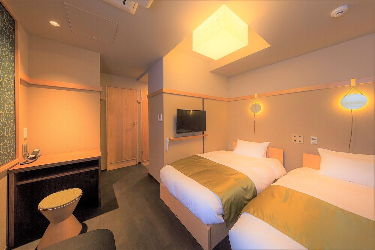 A hotel room with two beds, a TV, a desk with a chair, and a desk lamp.