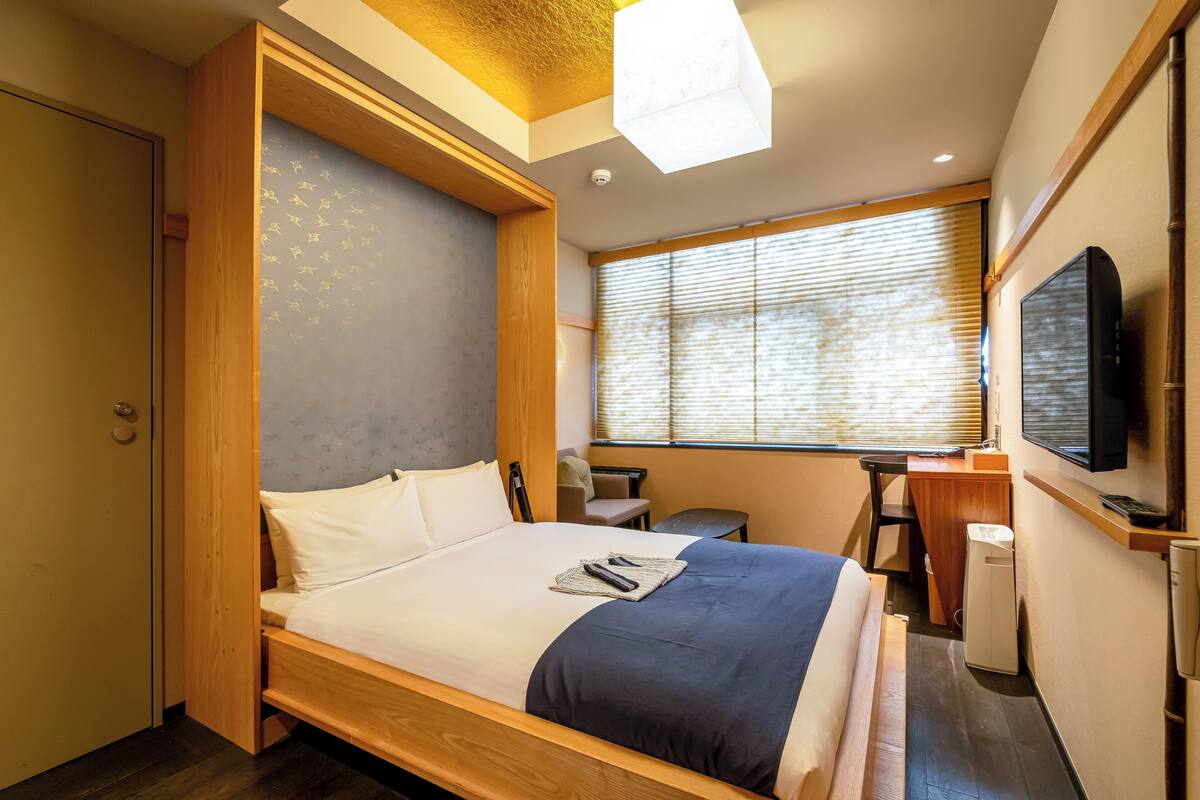 A compact hotel room with a bed, a desk, a chair, a television, and a window with blinds.