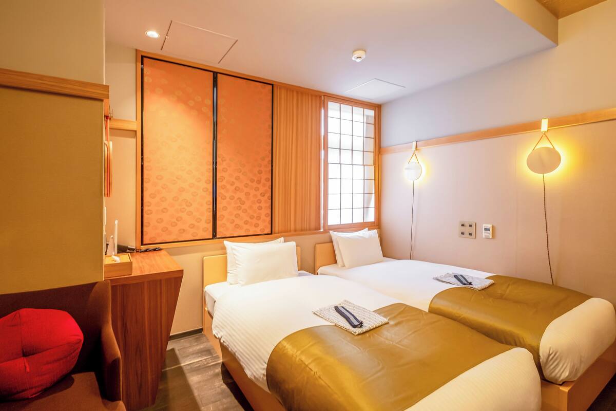 A hotel room with two beds, a desk, a red chair, and a sliding door.