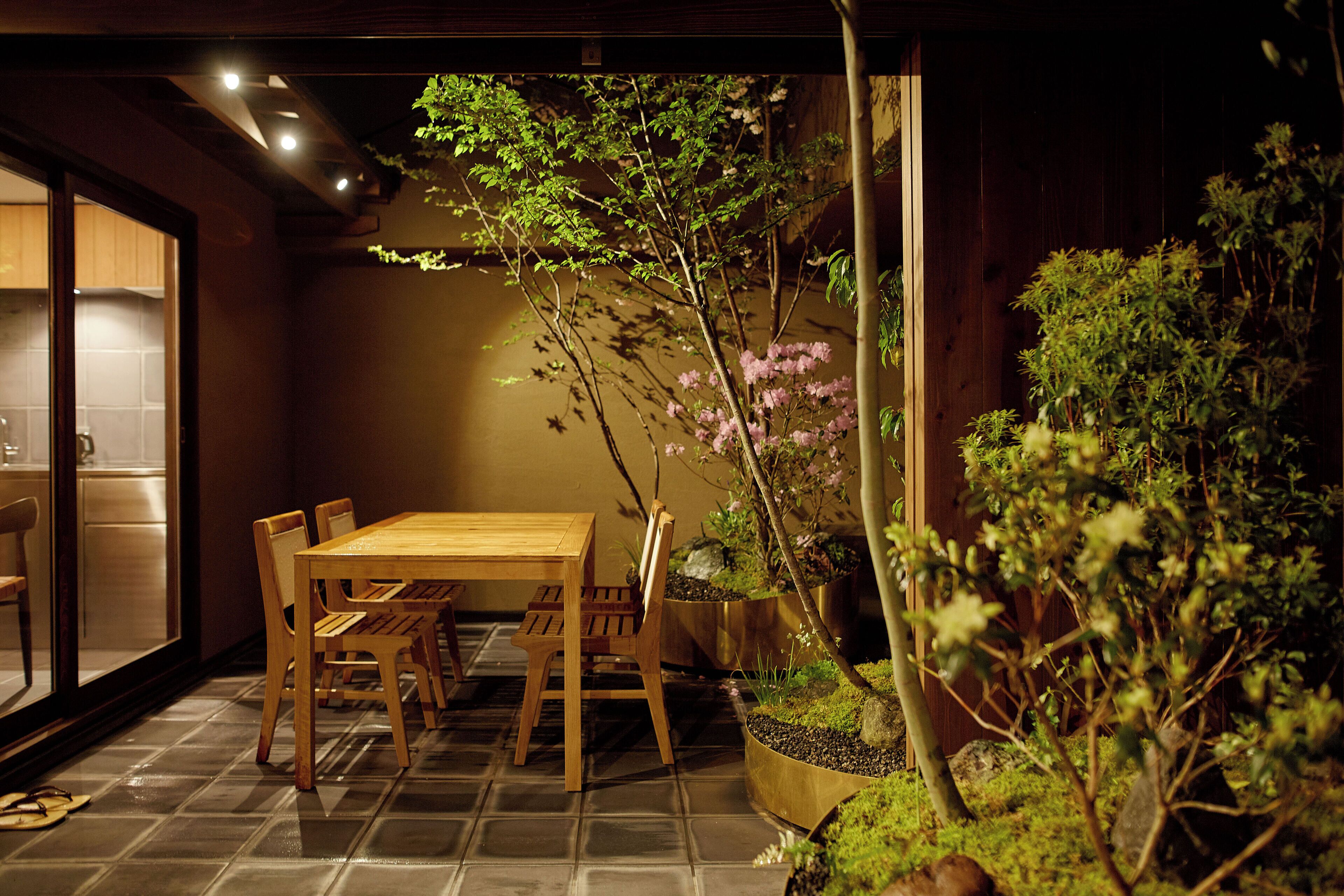 Un coin repas avec une table et des chaises en bois, entouré de plantes en pot et d’un petit arbre aux fleurs roses.