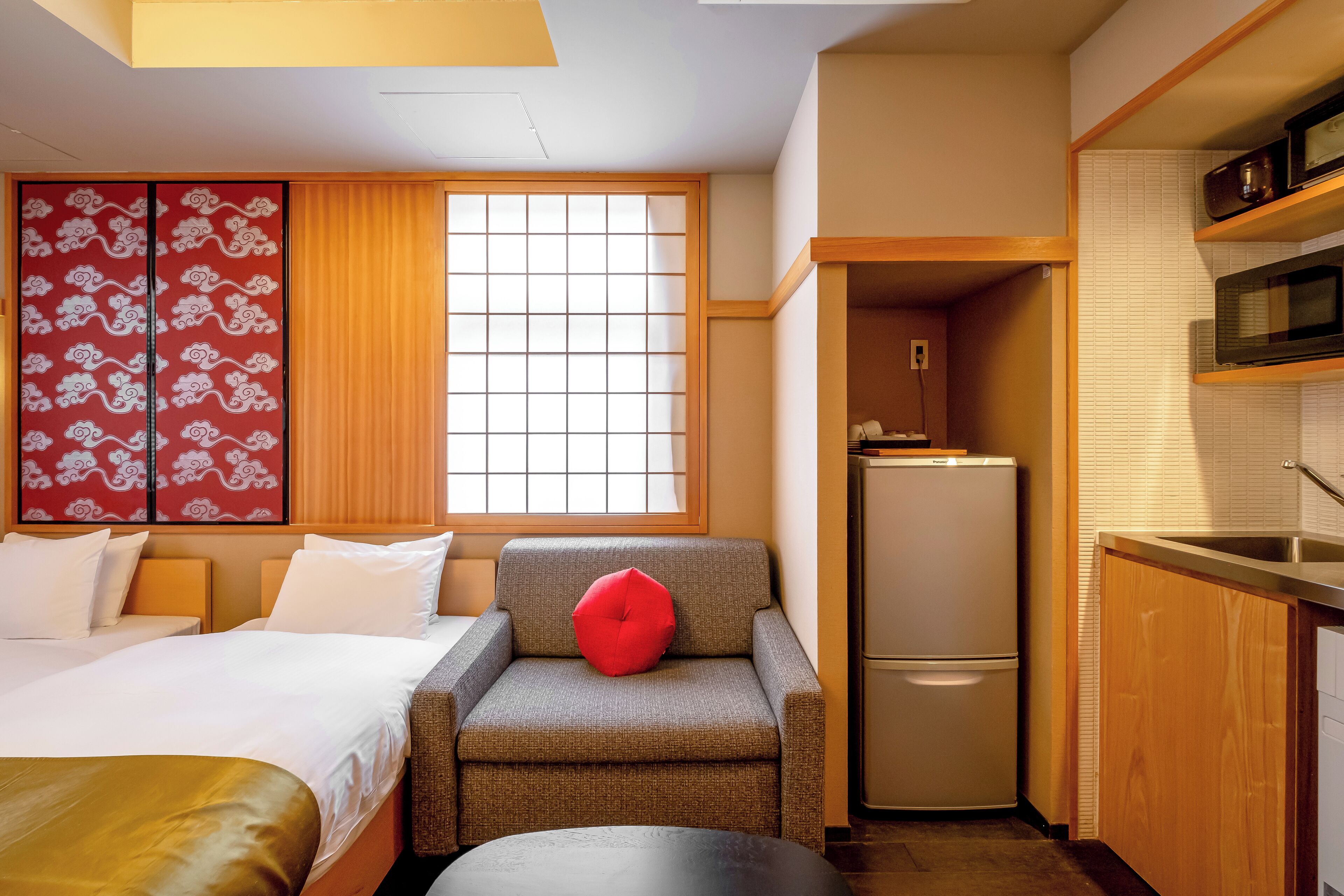 A compact hotel room with a bed, a small sofa with a red cushion, a kitchenette with a refrigerator, and a window with a grid pattern.