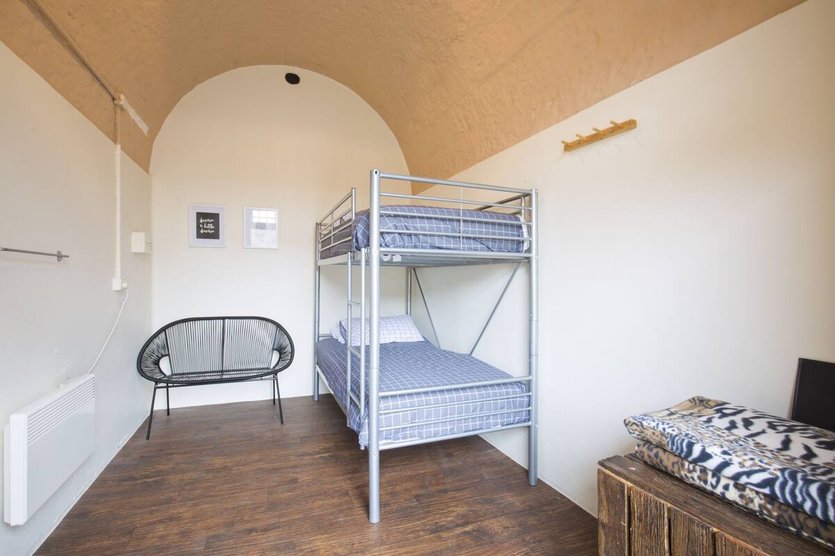 Een stapelbed met een geruit matras, een zwarte stoel en een klein houten tafeltje in een kamer met houten vloeren en witte muren.