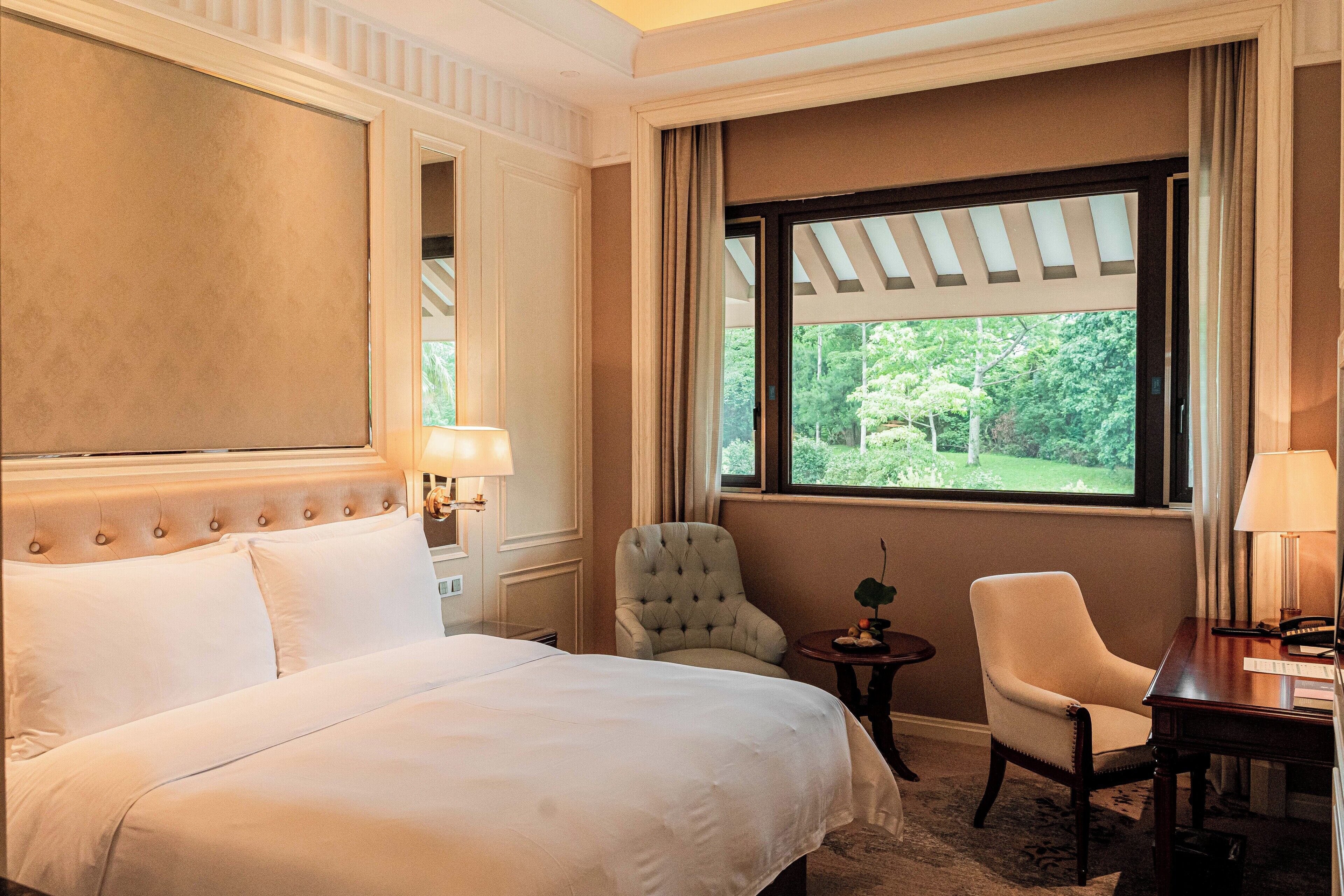 A hotel room with a large bed, two chairs, a small table, and a window with a view of greenery.