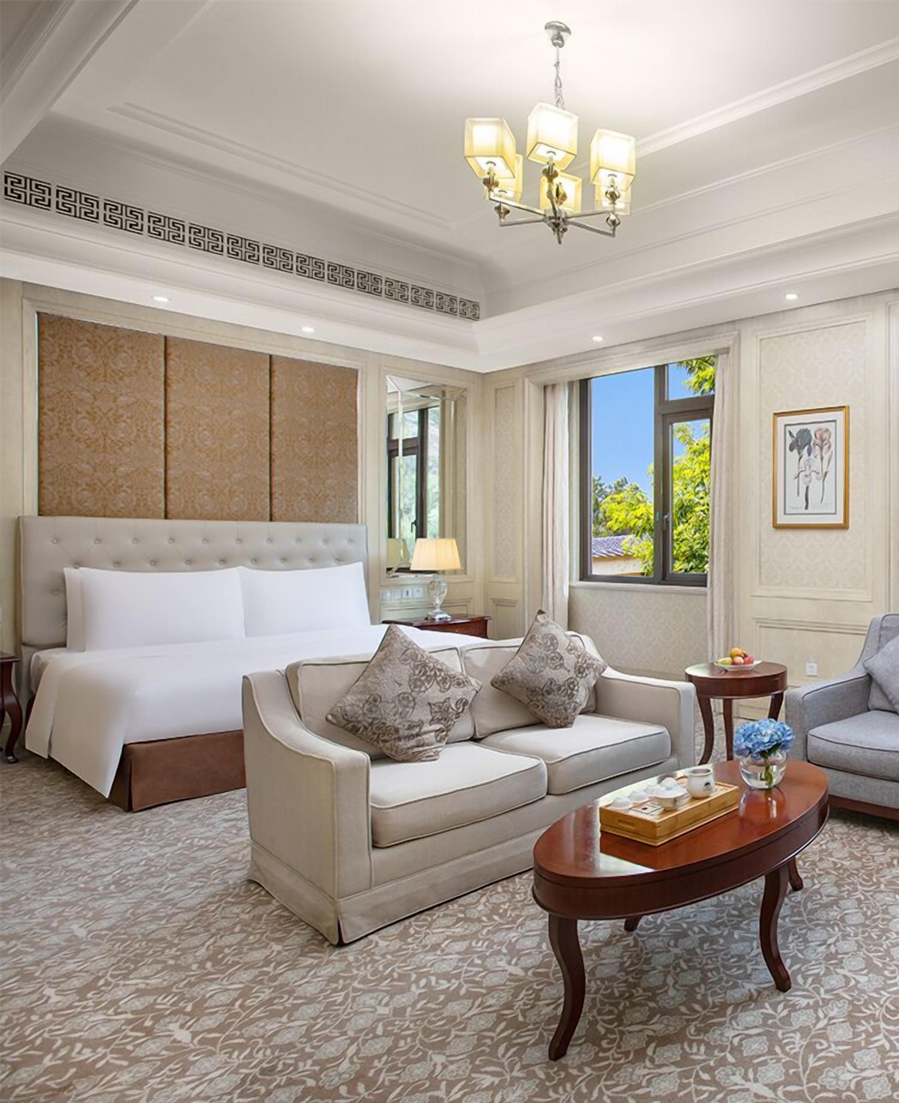 A hotel room with a large bed, a sofa, a coffee table, and a view of greenery outside.
