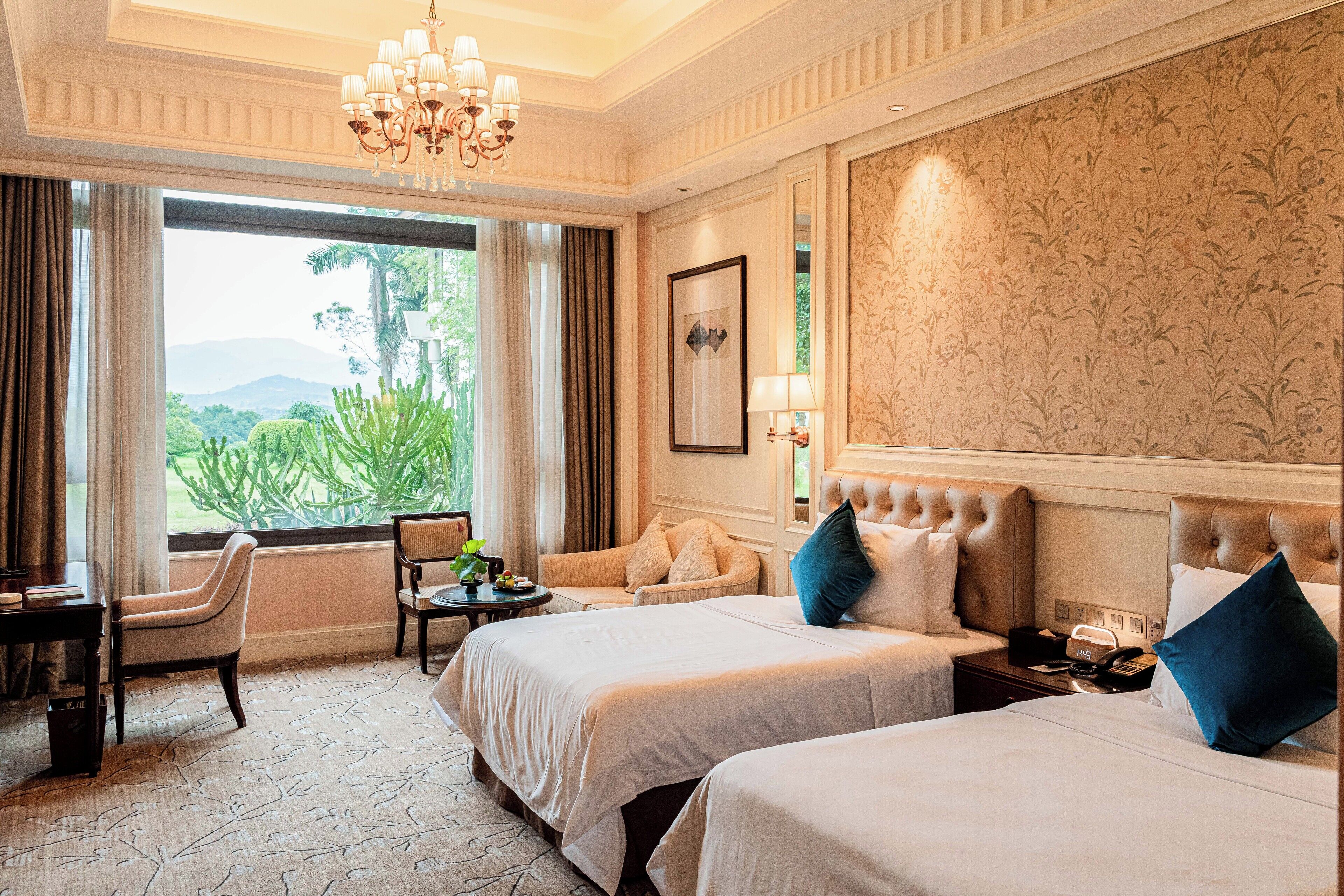 A hotel room with two beds, a desk, a chair, and a view of greenery outside.
