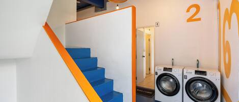Un área de lavandería con una escalera azul, dos lavadoras y un letrero que indica el baño de mujeres.