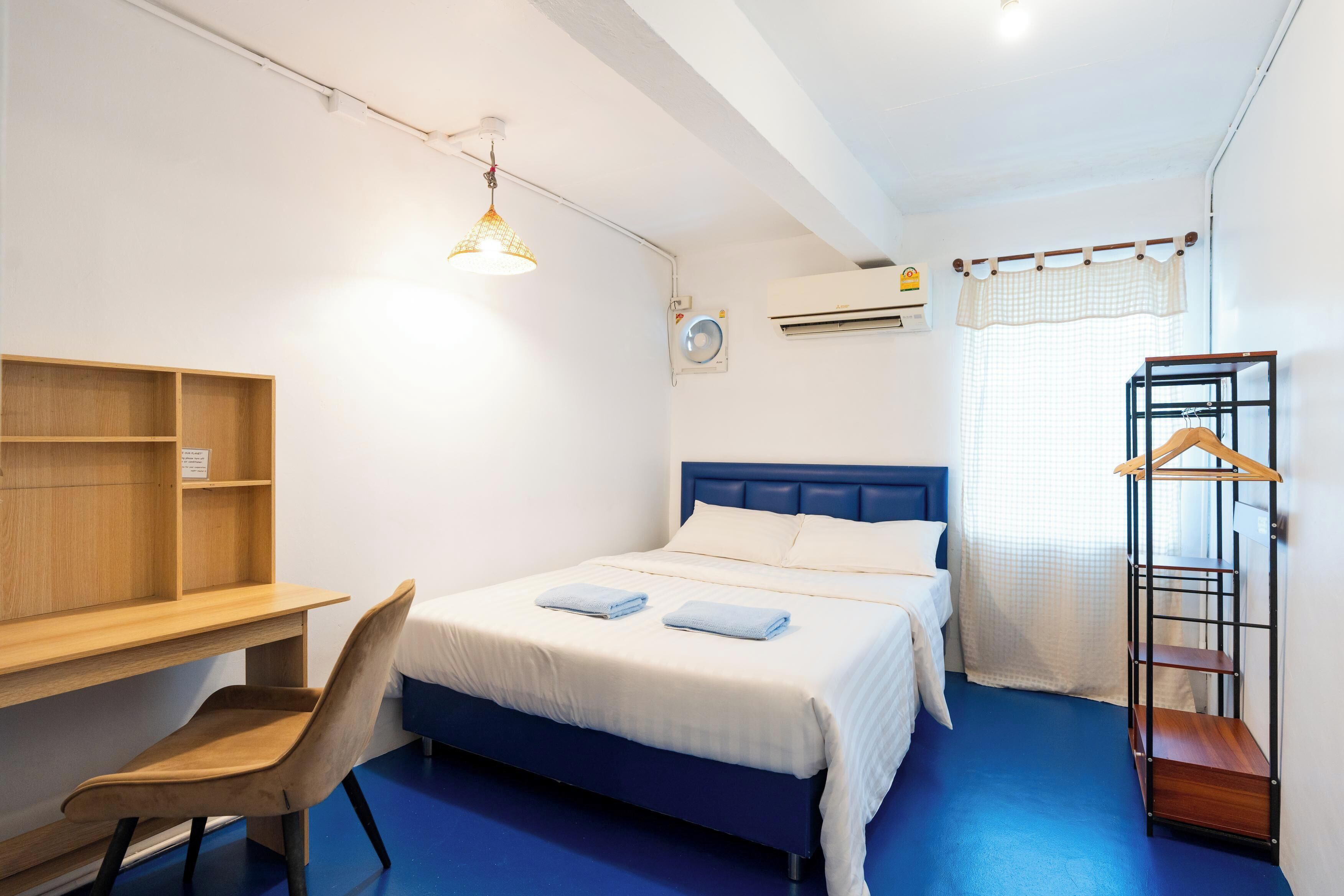 A room with a bed, a desk, a chair, a hanging light fixture, an air conditioning unit, and a window with a curtain.