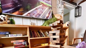 A wooden Jenga tower on a table with scattered blocks, a television displaying a cityscape, and shelves with books and board games. - Arawana Hostel Silom (Bangkok)