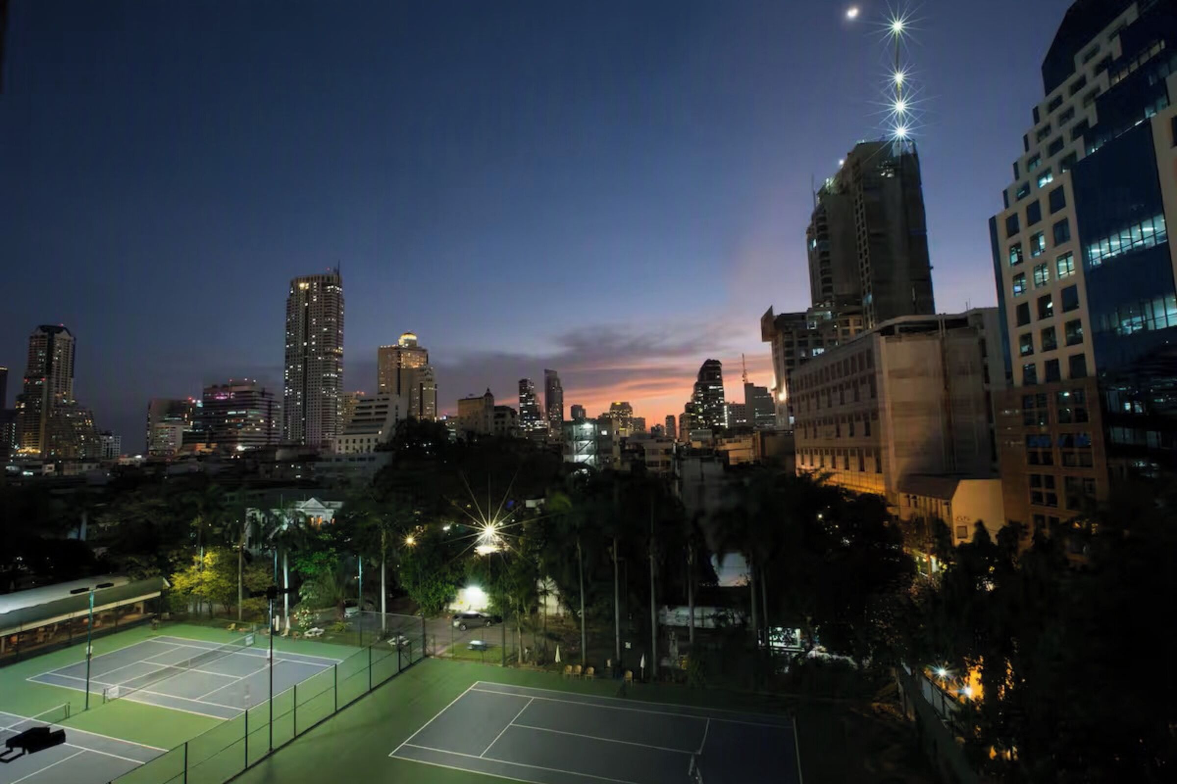A tennis court surrounded by urban buildings at dusk.