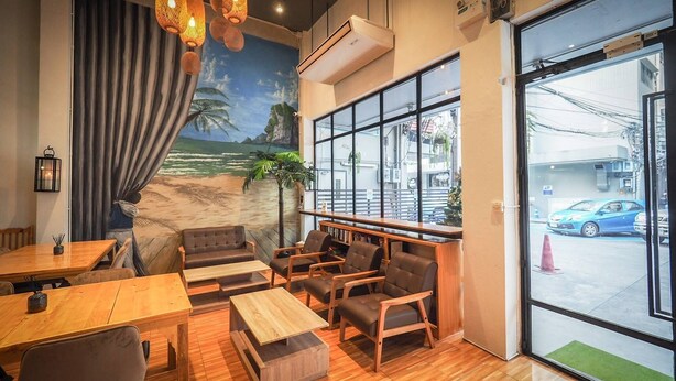 A cozy cafe with wooden furniture, a mural of a beach scene, and large windows offering a view of the street. - Arawana Hostel Silom (Bangkok)