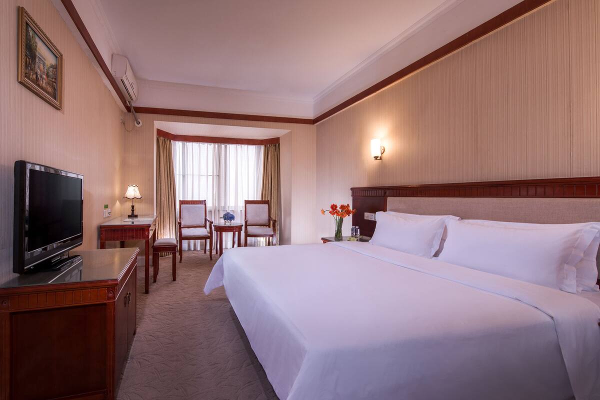 Une chambre d’hôtel avec un grand lit, un téléviseur posé sur une commode en bois, un bureau avec une lampe, une chaise et un vase de fleurs.