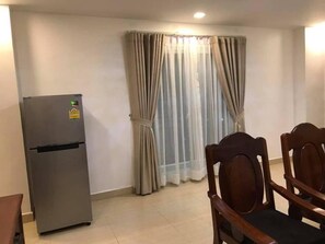 Un espacio habitable compacto con refrigerador, una mesa de comedor con sillas y una ventana con cortinas traslúcidas.