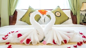 Una cama impecablemente hecha con pétalos de rosa y una decoración de toallas con forma de cisne, cortinas verdes y una ventana con vistas.
