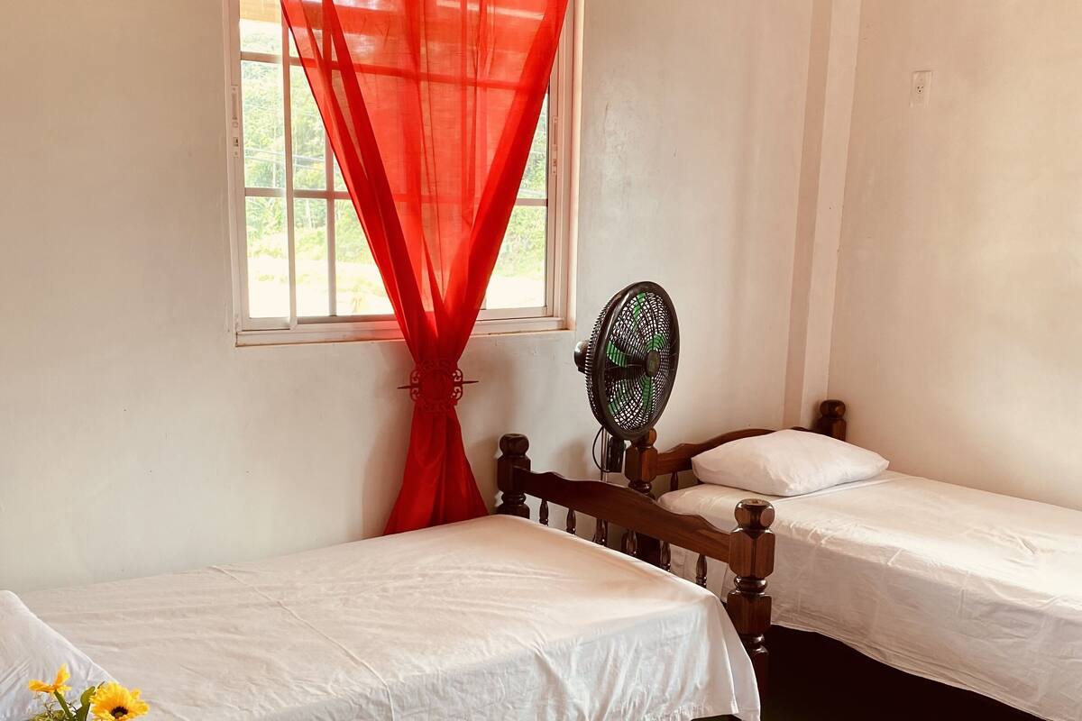 Una habitación con dos camas individuales, una mesita con mantel de encaje, un ventilador y una ventana con cortinas rojas.