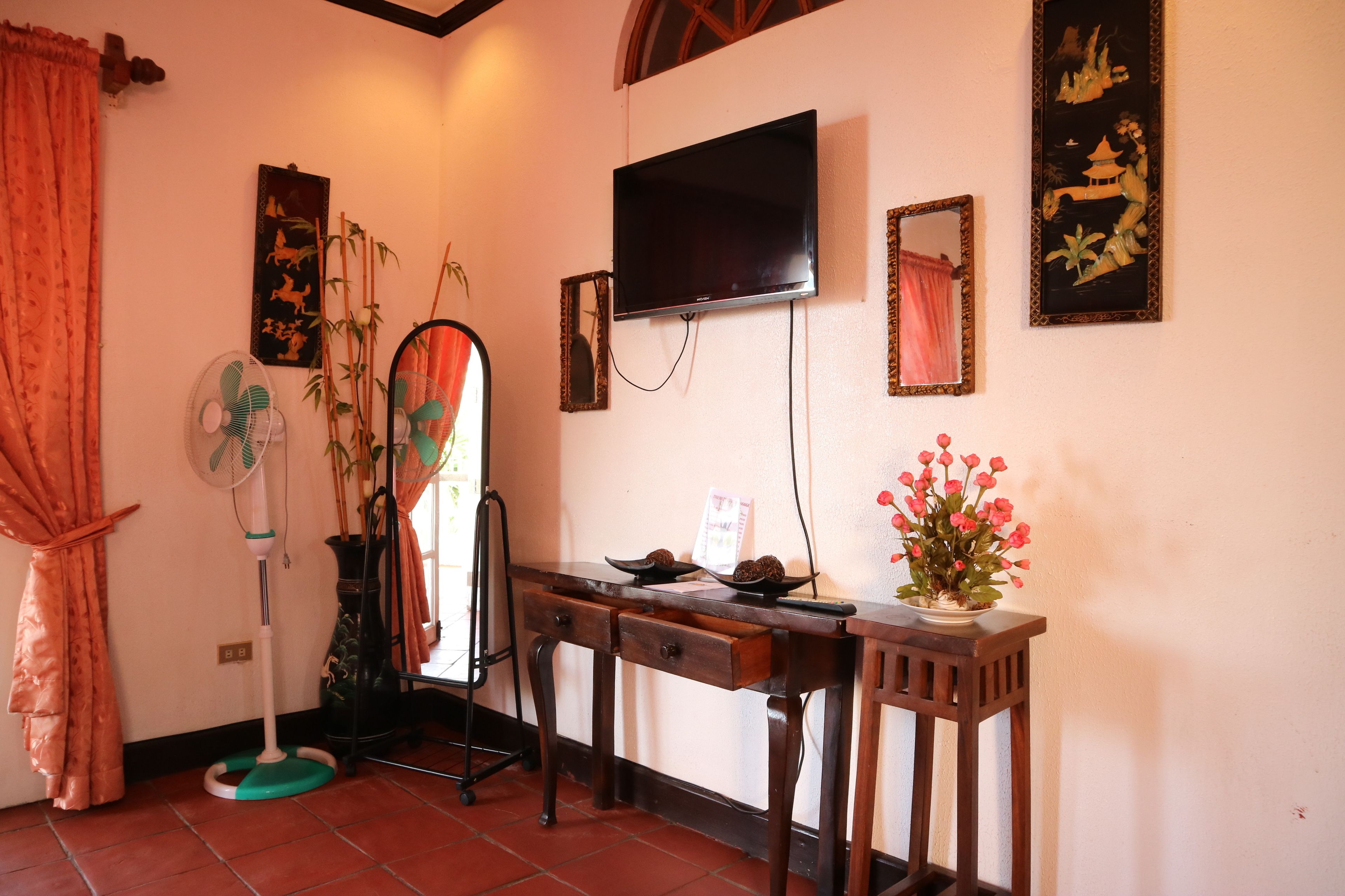 Una habitación con televisión montada en la pared, una mesa de madera con un jarrón de flores, un ventilador de pie y un espejo.