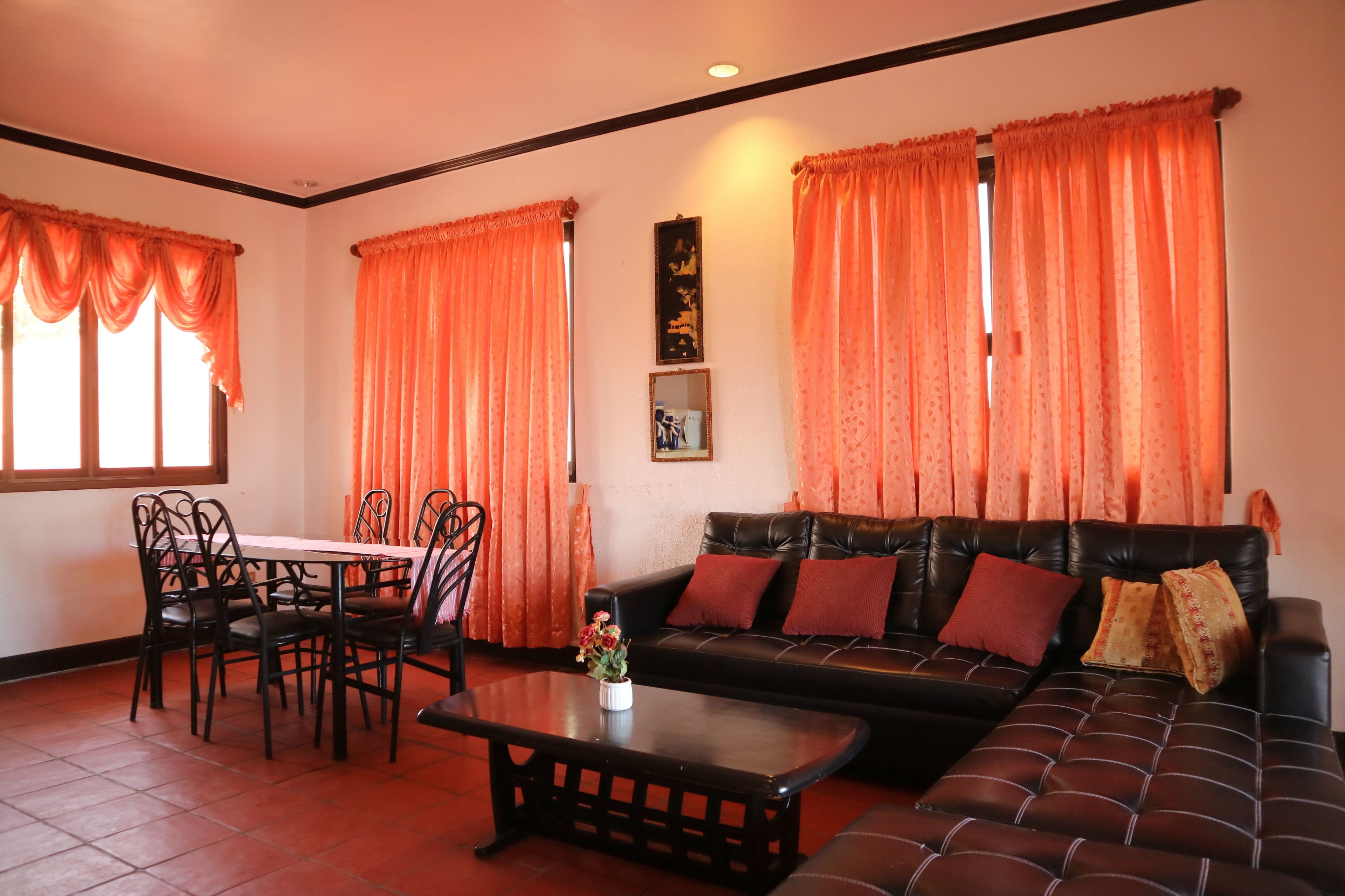 A living room with a black leather sofa, a dining table with chairs, and orange curtains.