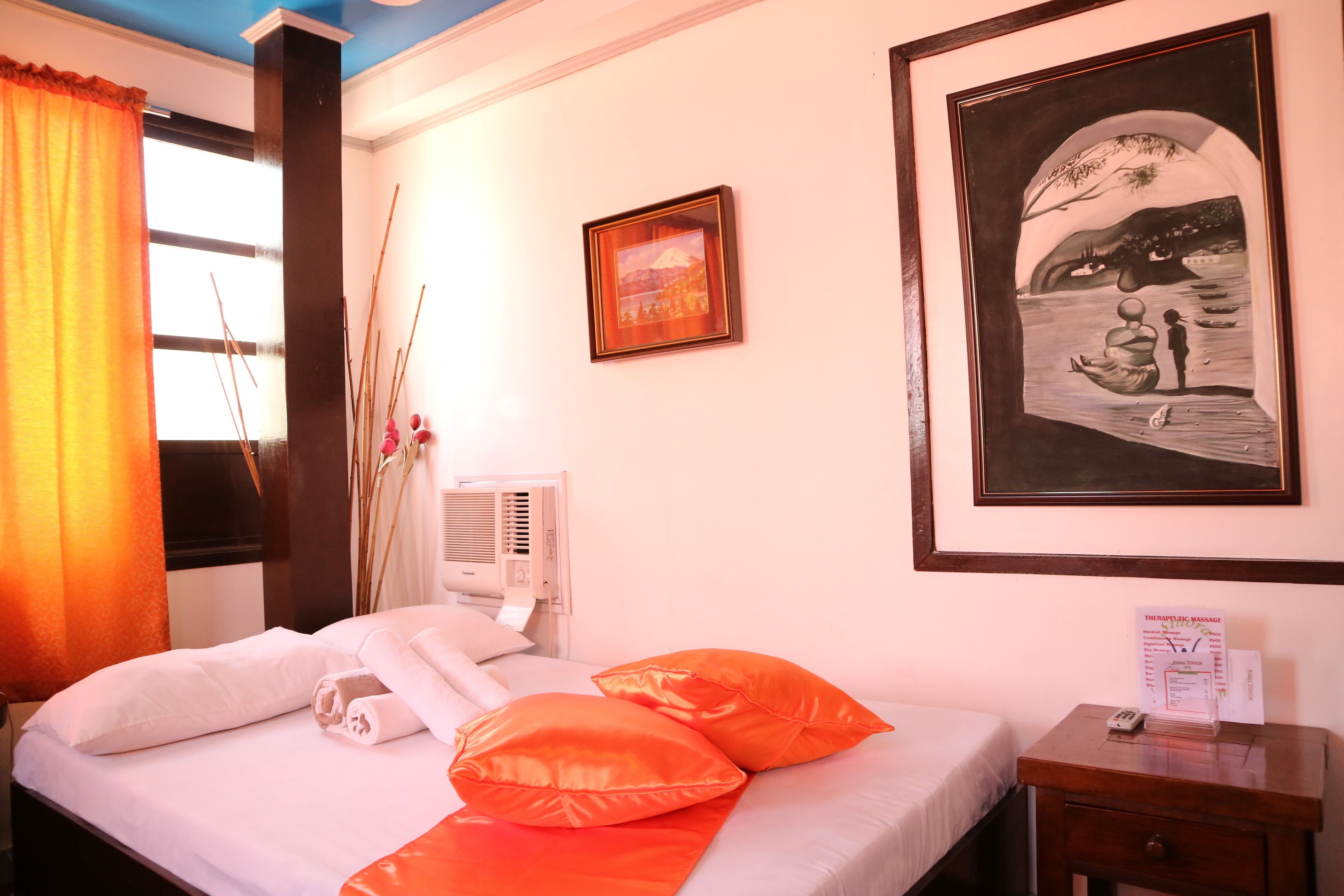 Habitación de hotel con cama, almohadones anaranjados, un cuadro enmarcado, una mesita de noche con un control remoto y una ventana con cortinas.