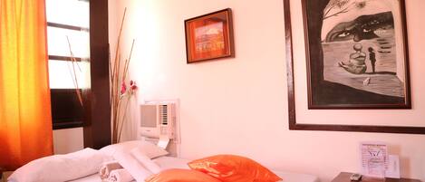 Une chambre d’hôtel avec un lit, des oreillers orange, un tableau encadré, une table de chevet avec une télécommande et une fenêtre avec des rideaux.