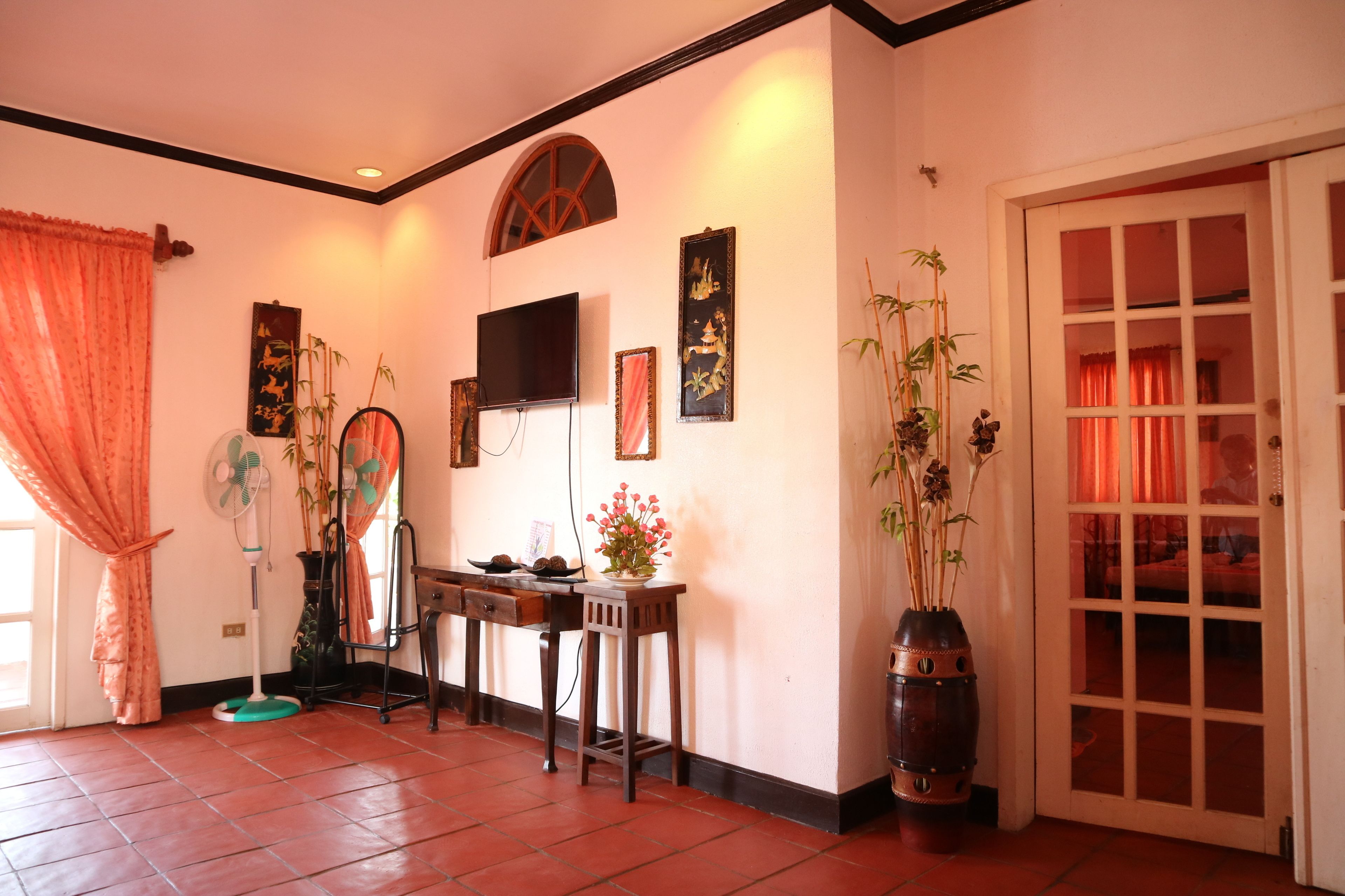 Una habitación tradicional con suelo de baldosas, un televisor montado en la pared, una mesa de madera con un florero y un amplio ventanal con cortinas.