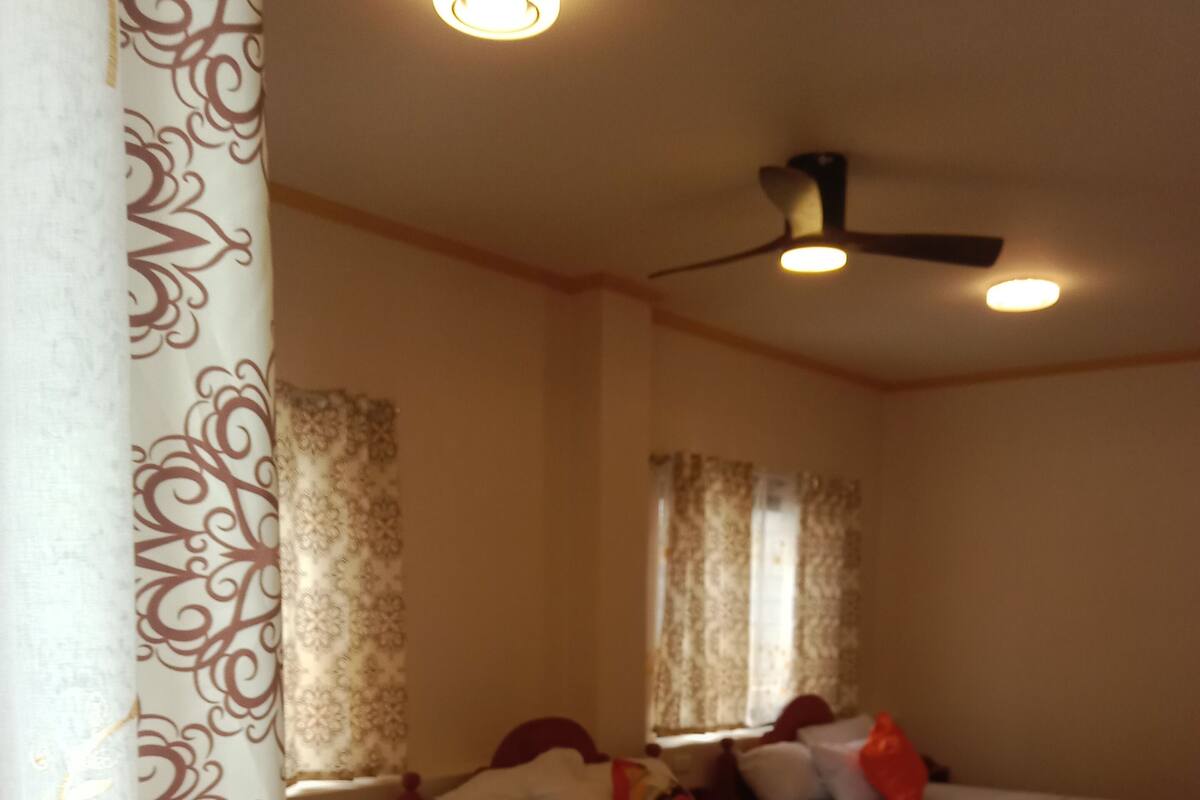 Une chambre d’hôtel avec trois lits simples, chacun recouvert d’une couverture blanche et orange, et dotée d’un ventilateur de plafond.
