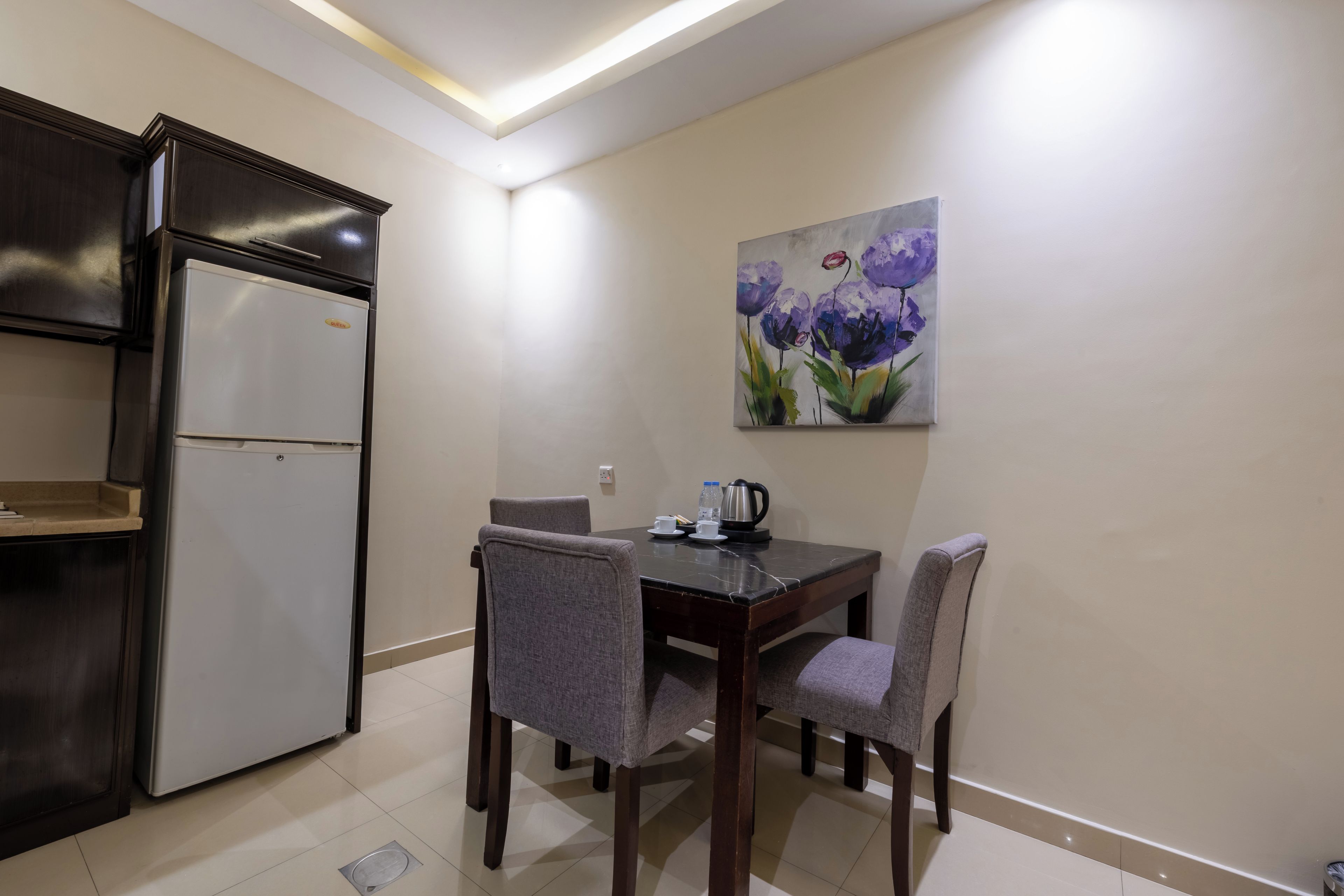 A compact kitchen with a refrigerator, a dining table with four chairs, and a painting on the wall.