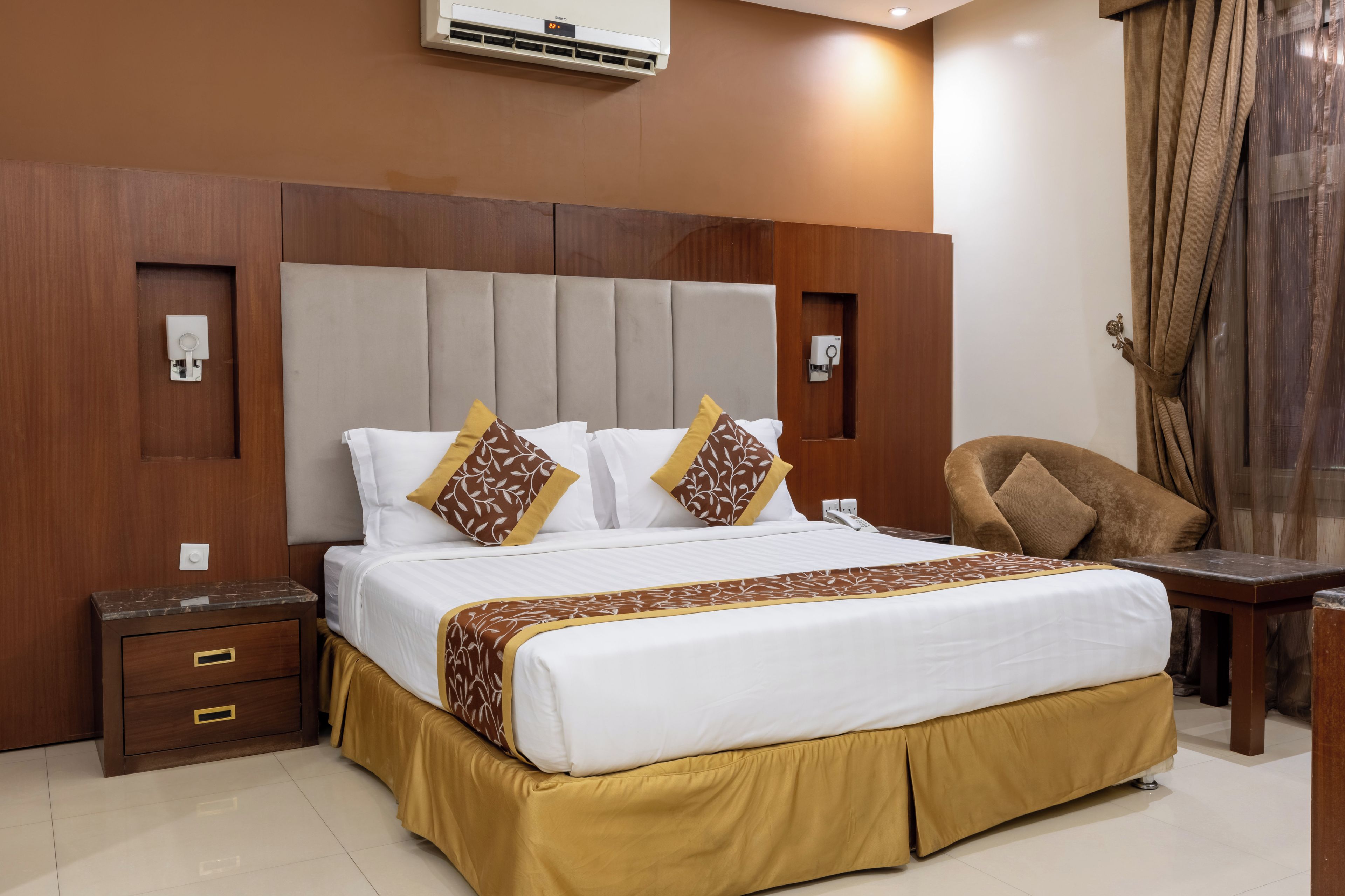 A hotel room with a large bed, bedside tables, a desk, and an air conditioning unit on the wall.