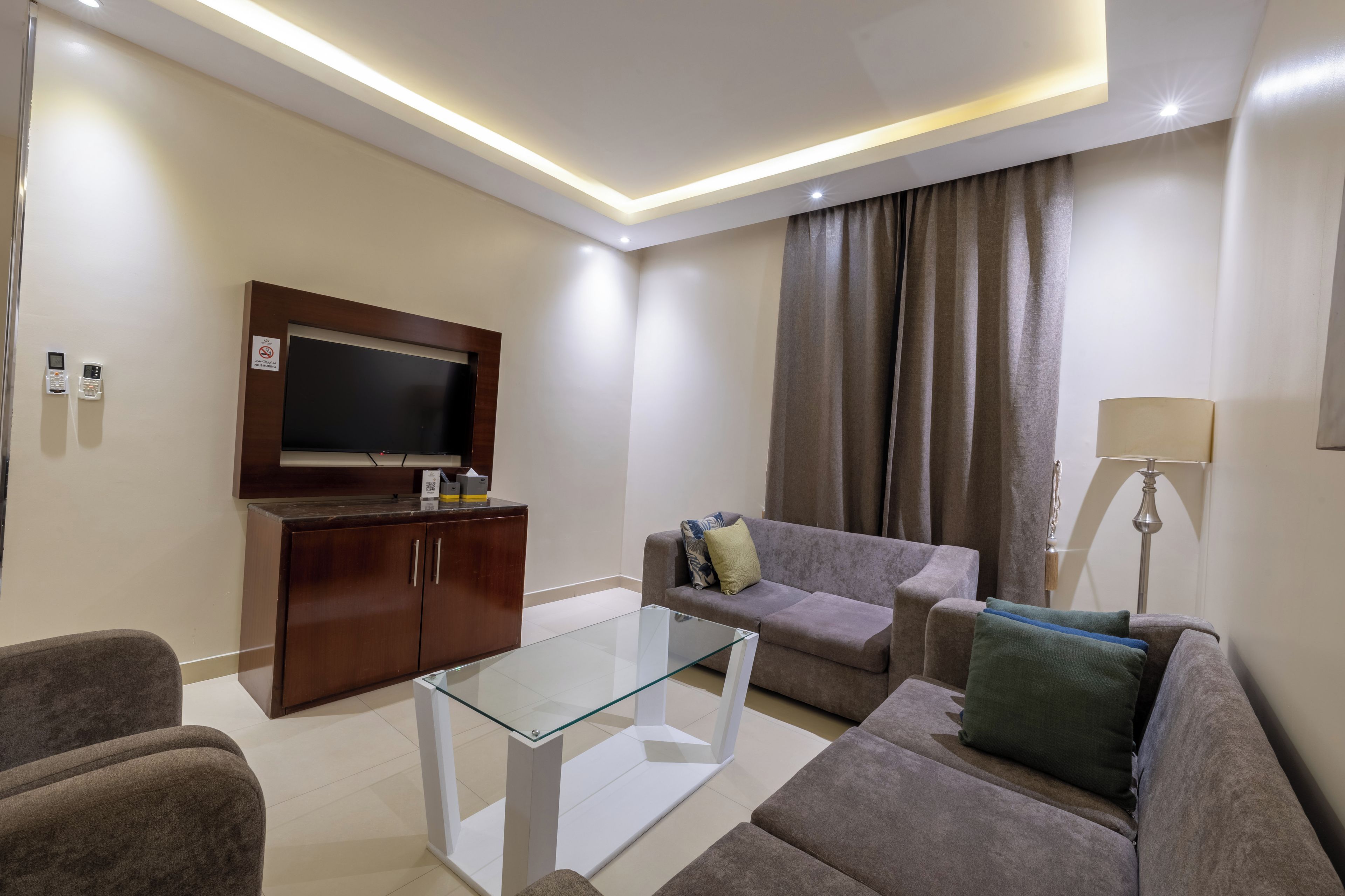 A modern living room with a flat-screen TV, wooden cabinet, glass coffee table, grey sofa, and a lamp.