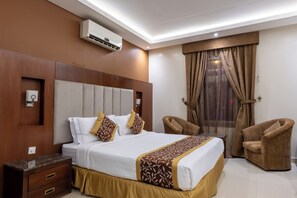 A hotel room with a large bed, two armchairs, a wooden headboard, an air conditioning unit, and a window with curtains.