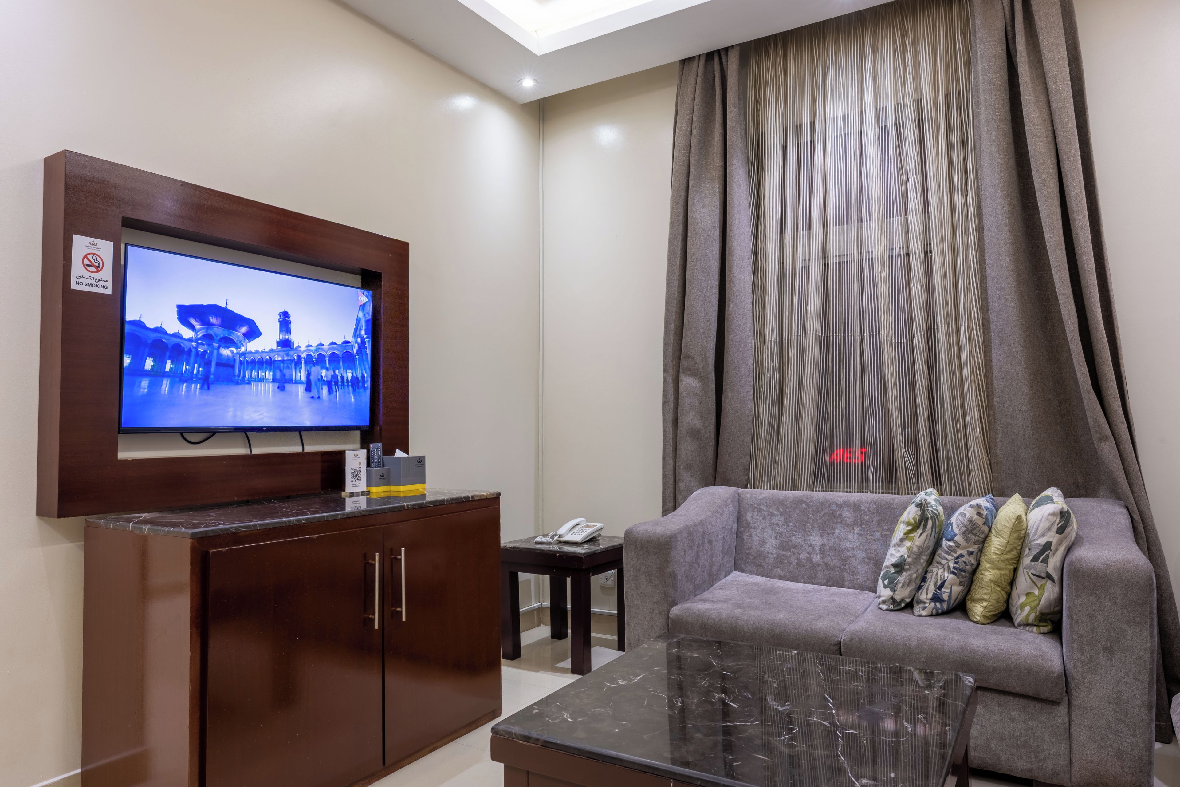 A hotel room with a television mounted on a wooden cabinet, a grey sofa with patterned cushions, a small table with a telephone, and a curtain with a vertical striped pattern.
