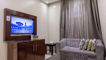 A hotel room with a television mounted on a wooden cabinet, a grey sofa with patterned cushions, a small table with a telephone, and a curtain with a vertical striped pattern.