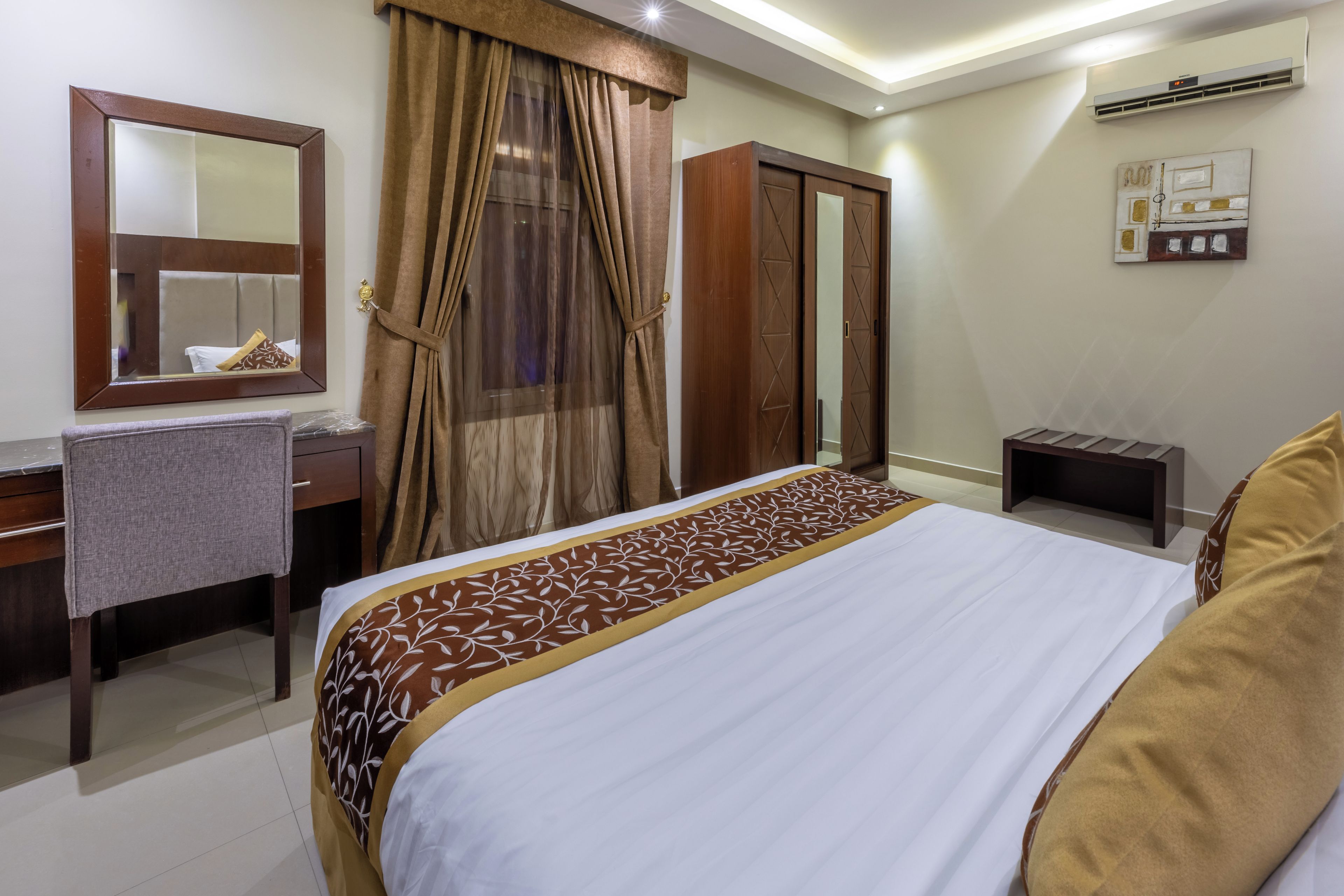 A hotel room with a bed, a desk, a mirror, a wardrobe, and an air conditioning unit.