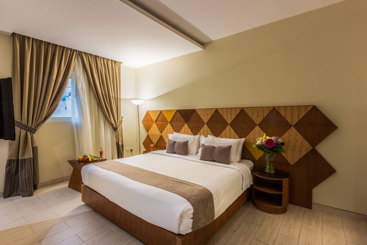 A hotel room with a large bed, wooden headboard, a nightstand with a vase of flowers, and a window with curtains.