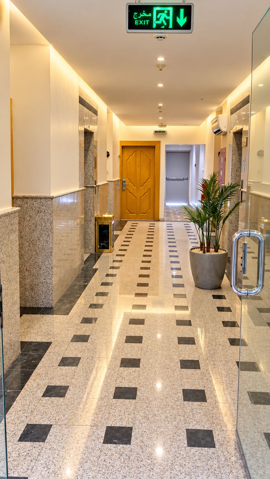 A hotel corridor with a polished tile floor, a potted plant, and an exit sign.
