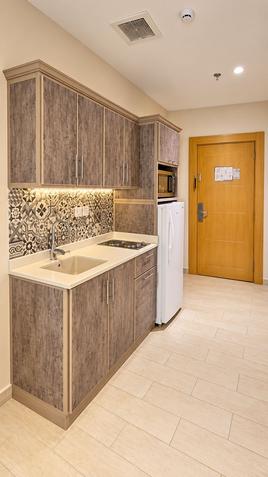 A compact kitchen with wooden cabinets, a microwave, a refrigerator, and a patterned backsplash.