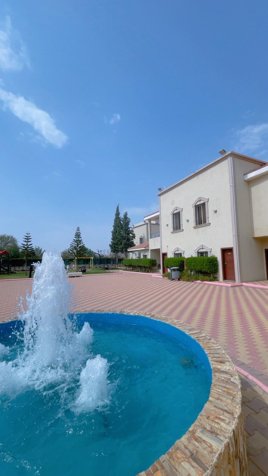 Une piscine avec une fontaine, entourée d’une zone pavée avec des carreaux décoratifs, et un bâtiment à la façade blanche et aux fenêtres.