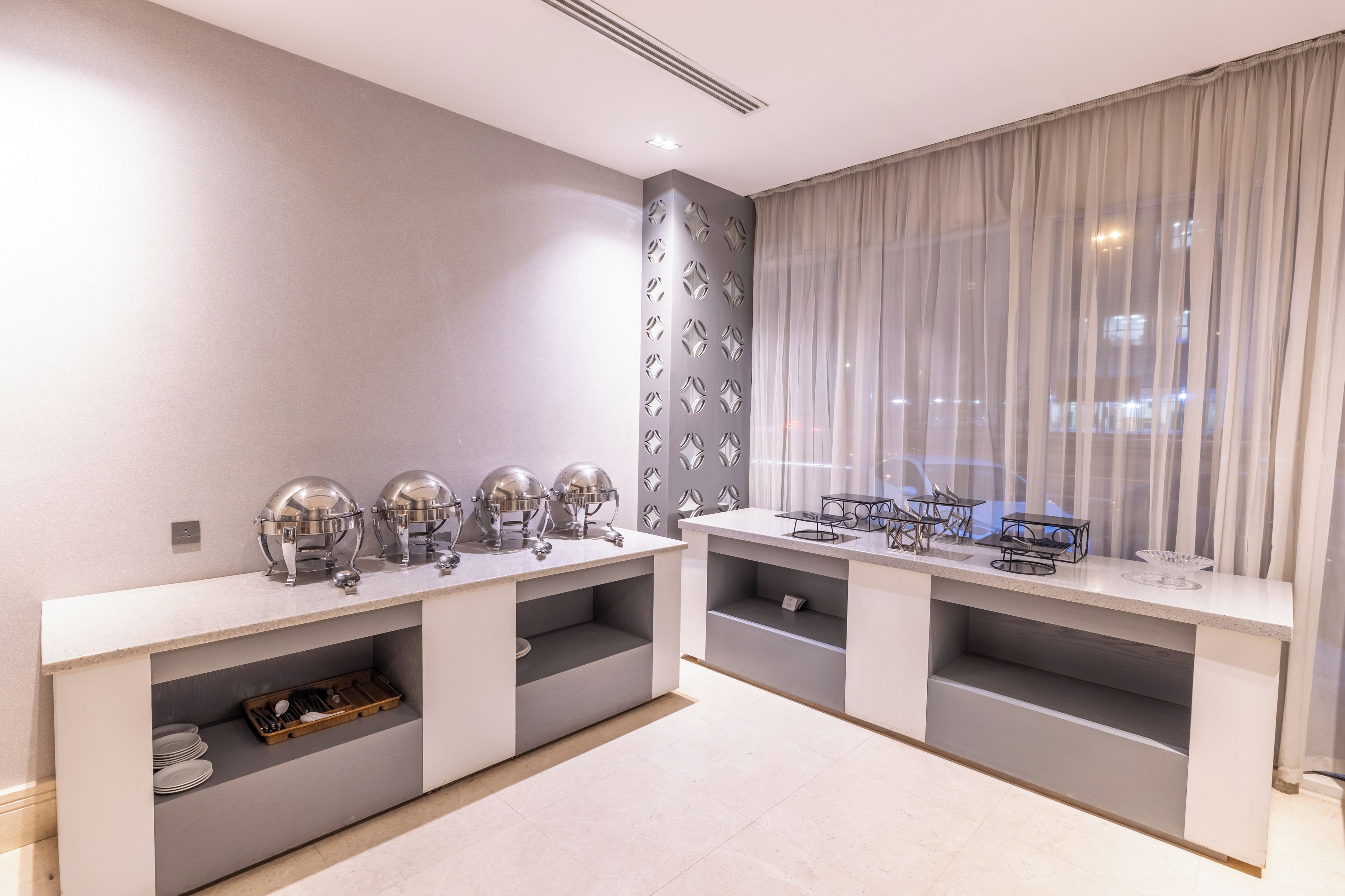 A modern room with two seating areas, each with a table and chairs. There are decorative wall panels and a large window with curtains.