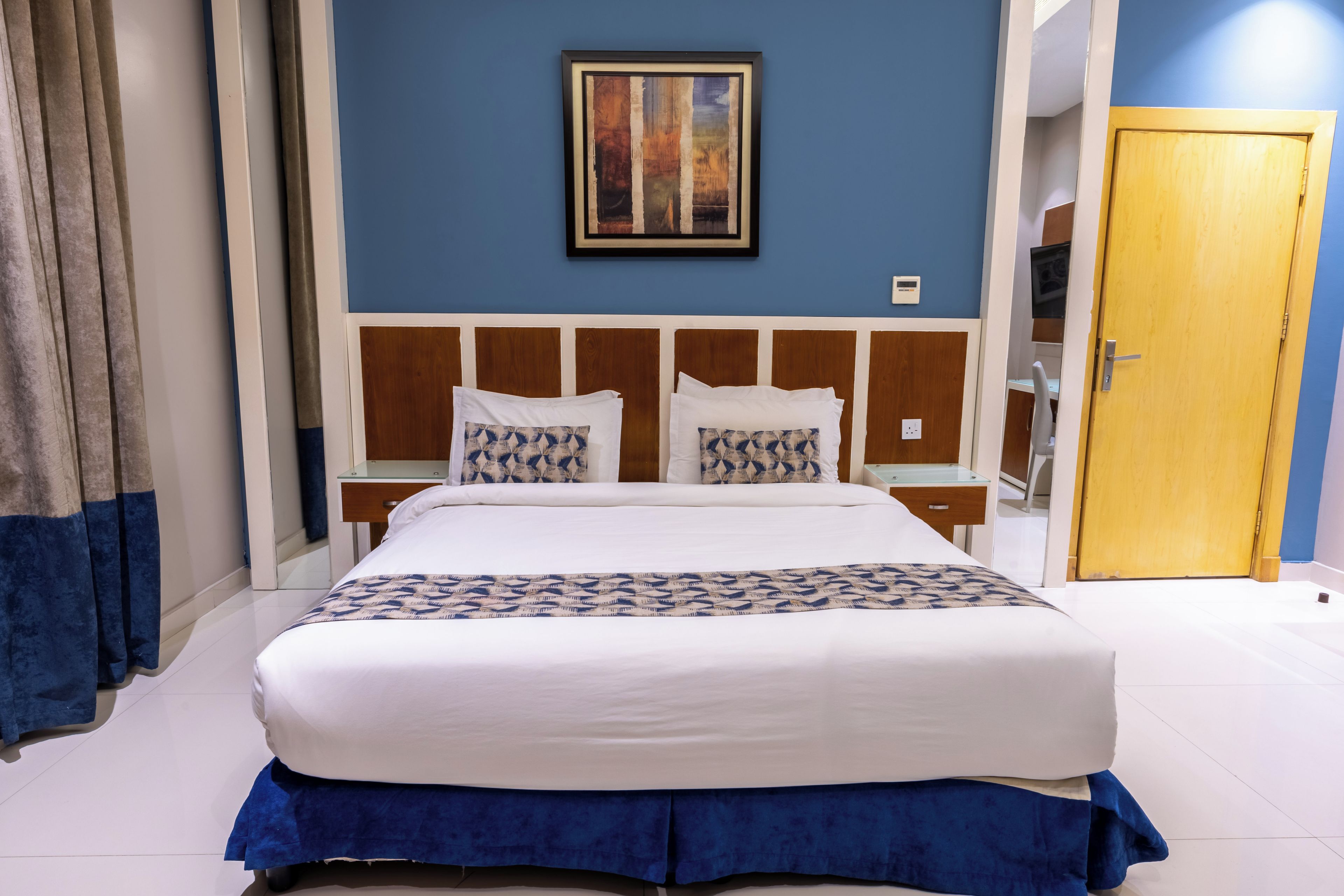 A hotel room with a large bed, a wooden headboard, a painting on the wall, and a door to the right.