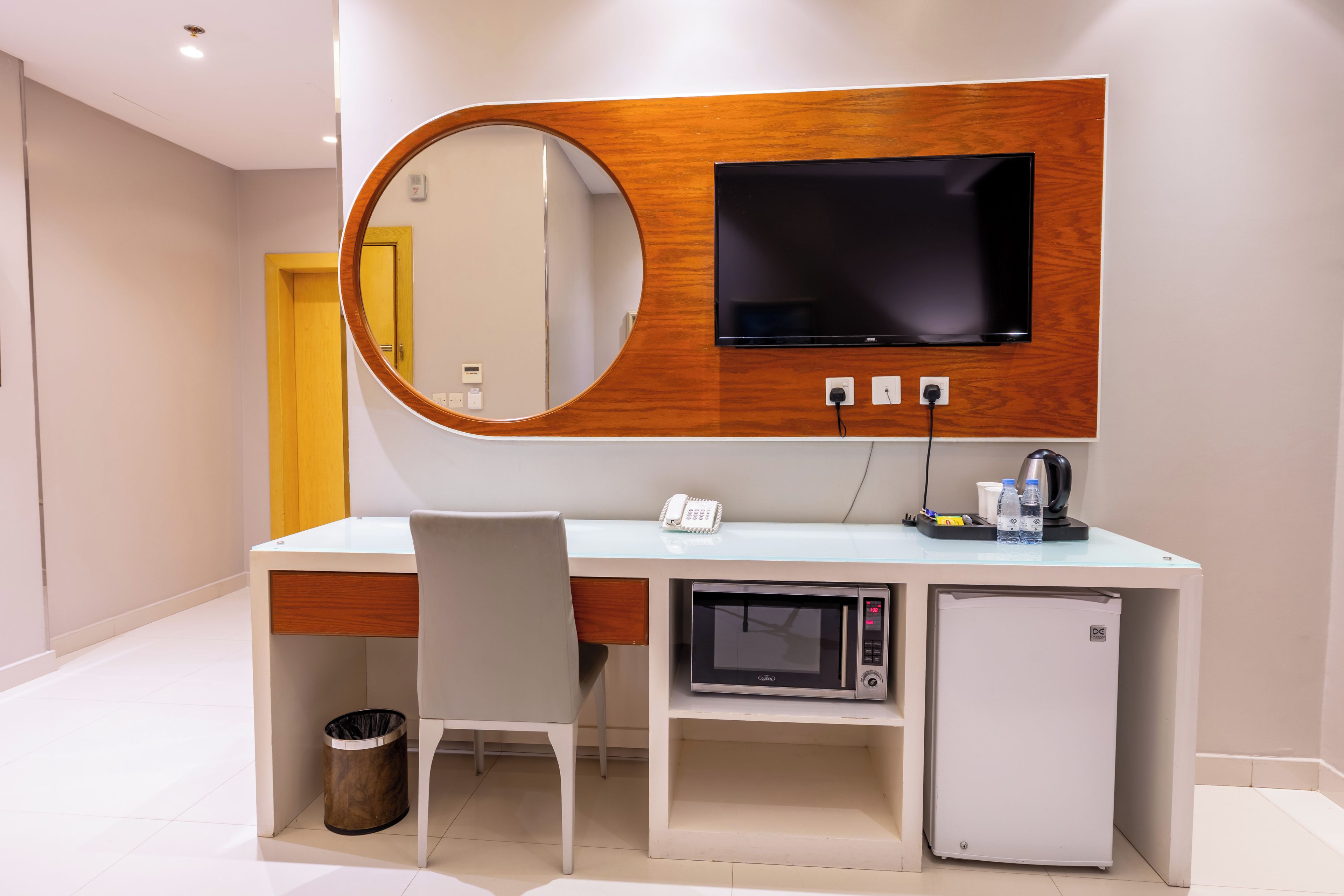 A modern hotel room with a wooden desk, a flat-screen TV, a microwave, a mini fridge, a trash bin, and a telephone.