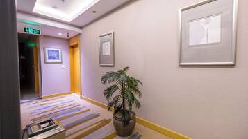 Un couloir d’hôtel avec un tapis à rayures, des œuvres d’art encadrées accrochées aux murs, une plante en pot et un panneau indiquant la sortie.