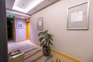 A hotel corridor with a striped carpet, framed artwork on the walls, a potted plant, and an exit sign.