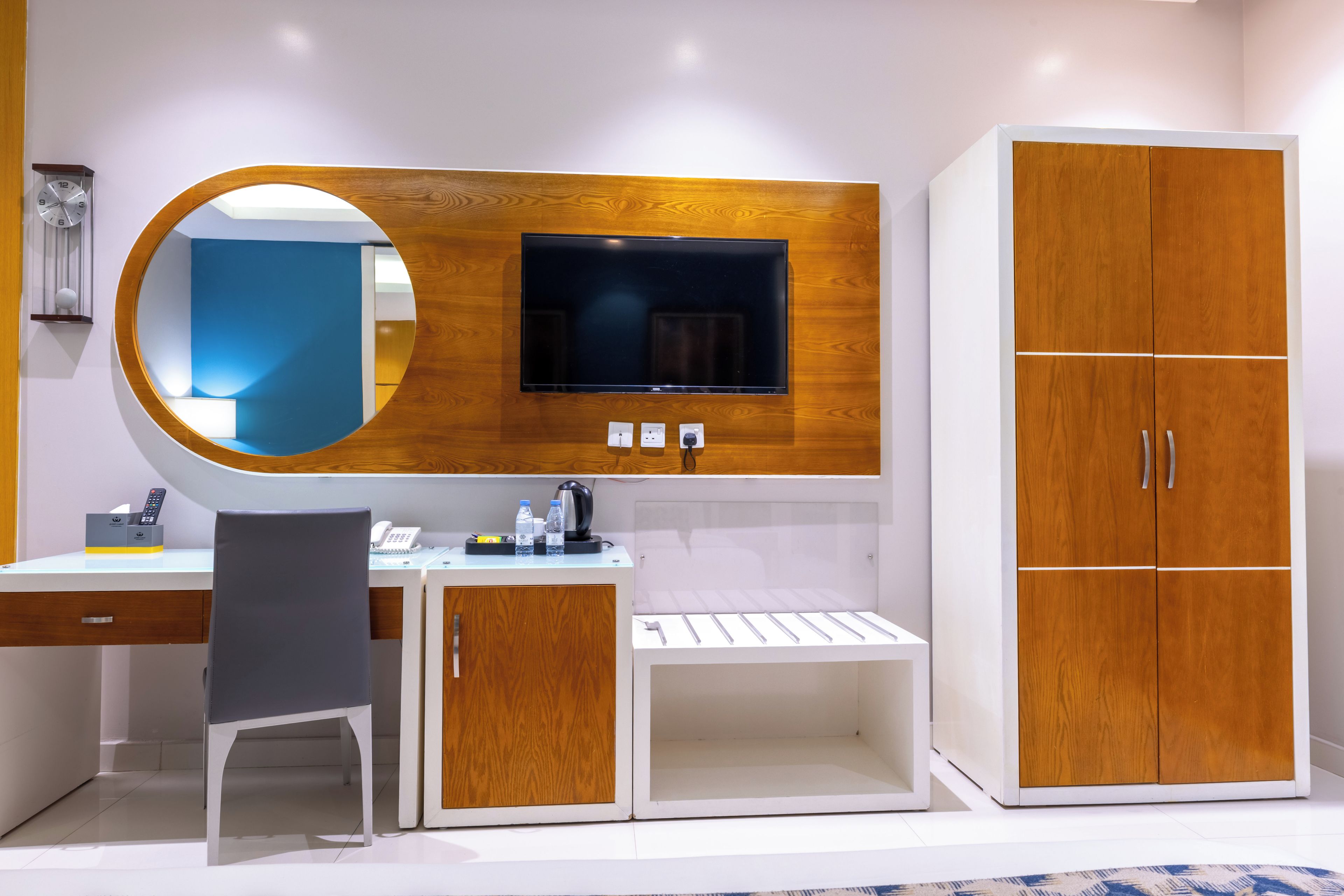 A modern hotel room with a wooden vanity, a built-in TV, a mirror, a desk with a chair, and a wardrobe.