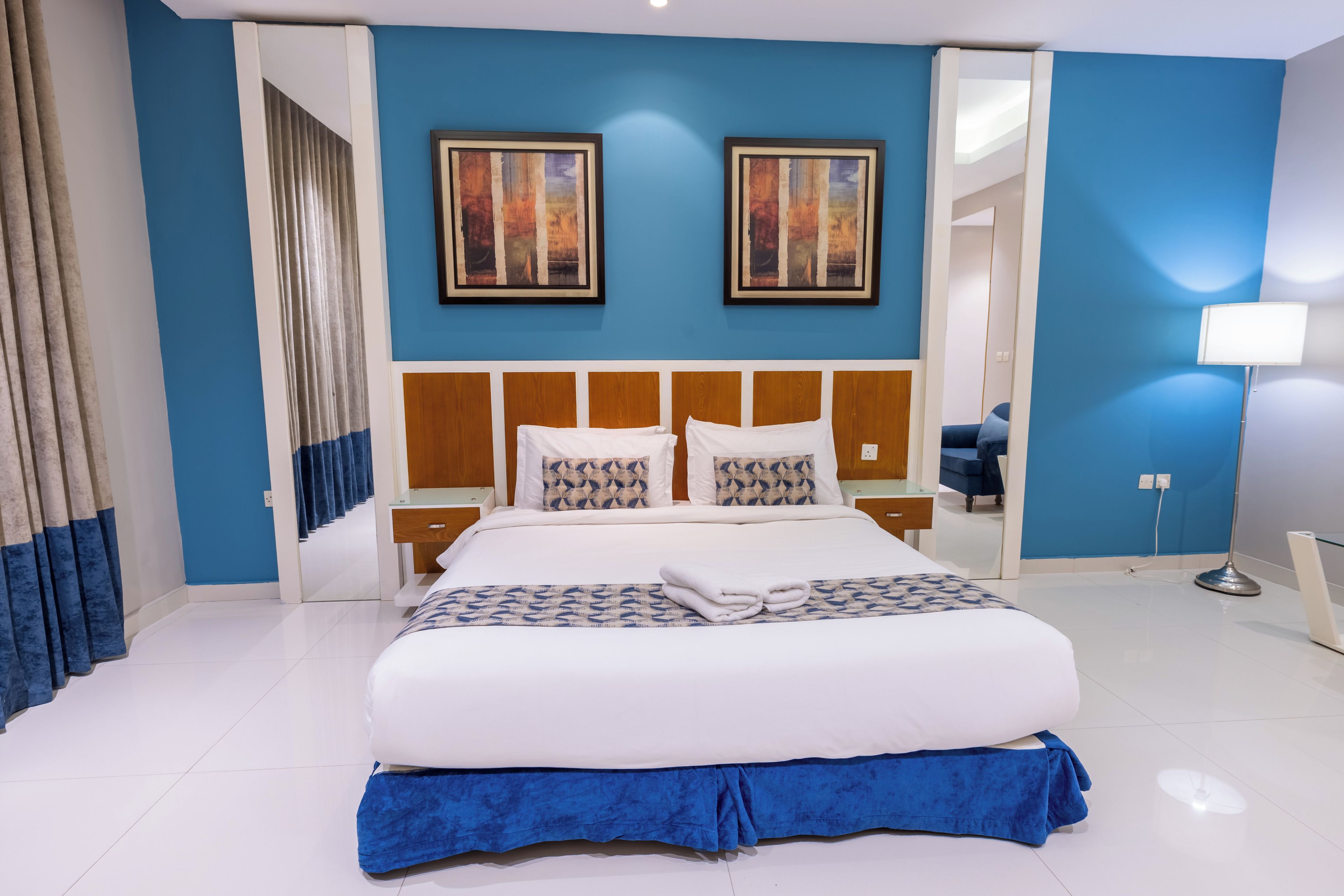 Quarto de hotel com uma cama grande, duas pinturas na parede, uma mesa de cabeceira, abajur e um espelho.