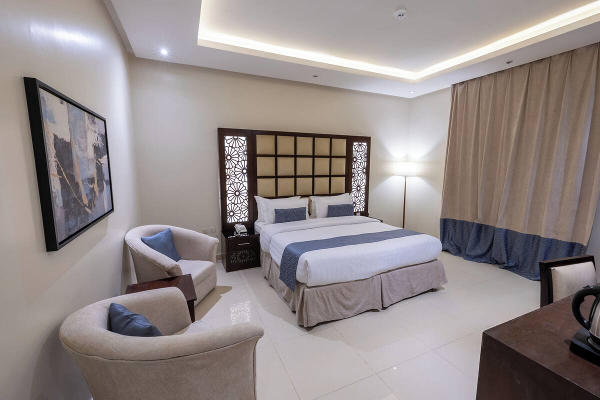 A modern hotel room with a large bed, a desk, a chair, and a bedside table.
